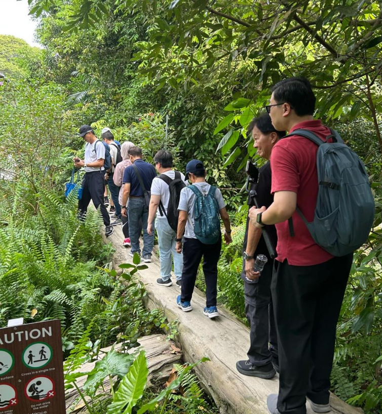 A group of people with backpacks walking single file on a narrow forest trail with a caution sign nearby.