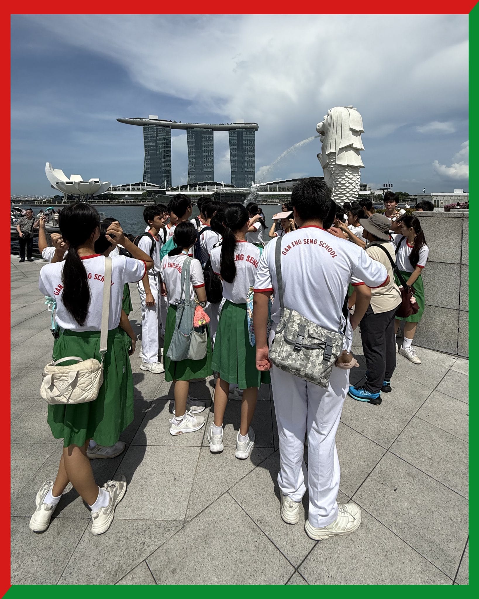 Gan Eng Seng School students in uniform stand near Singapore's Merlion statue with Marina Bay Sands behind.
