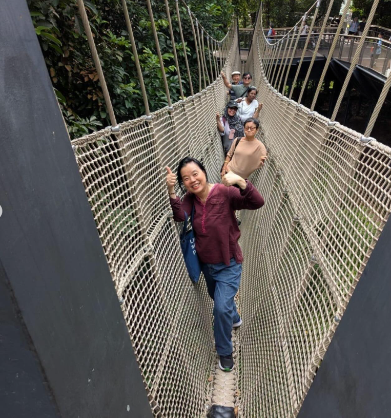 A group of people walking across a rope suspension bridge surrounded by green trees, with a woman giving thumbs up.