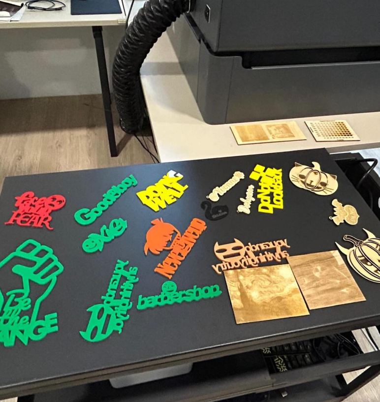 Colorful laser-cut word art pieces in red, green, yellow, and orange arranged on a black table near a laser cutter.