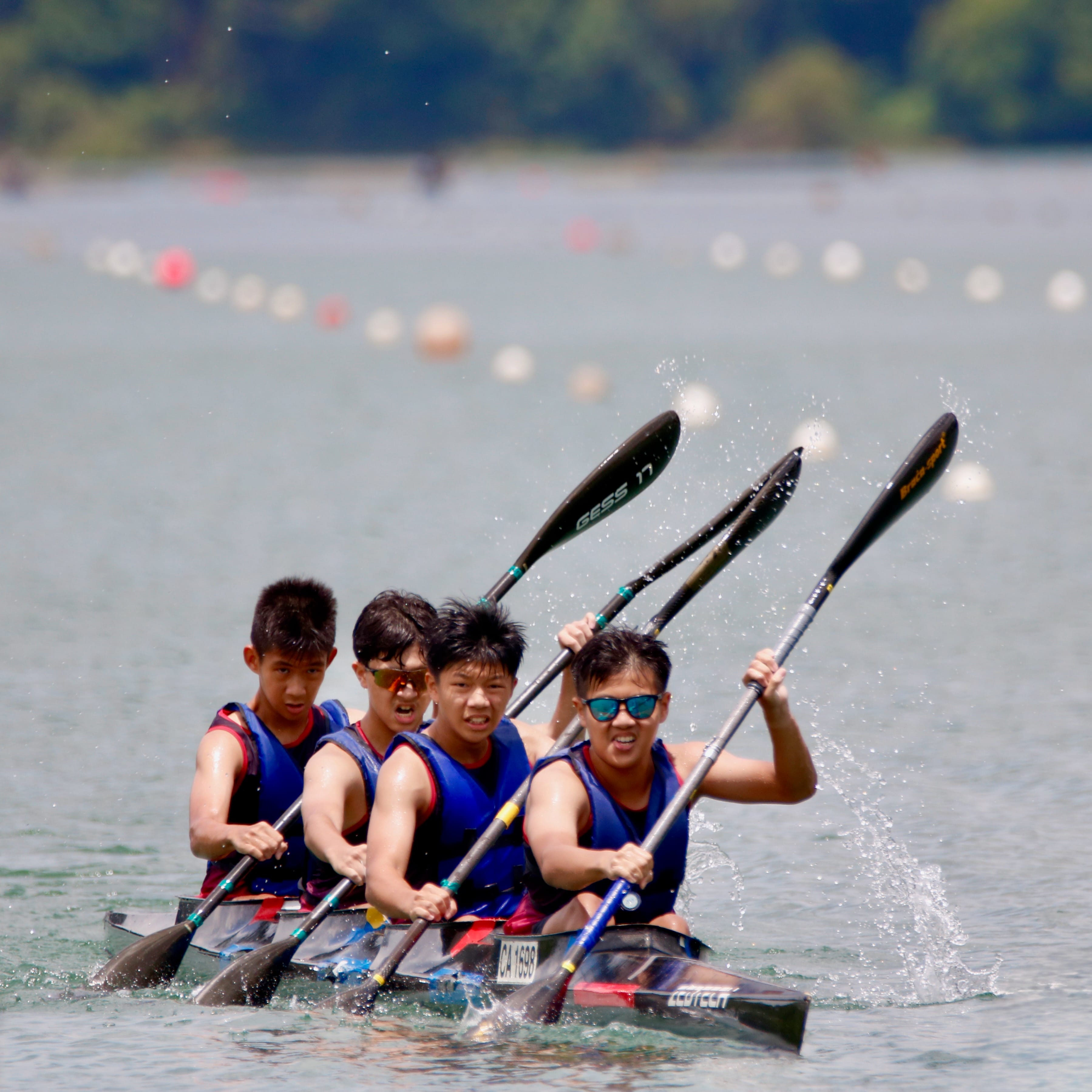 Four young boys in blue vests paddle kayaks on a lake with race buoys visible in the background.