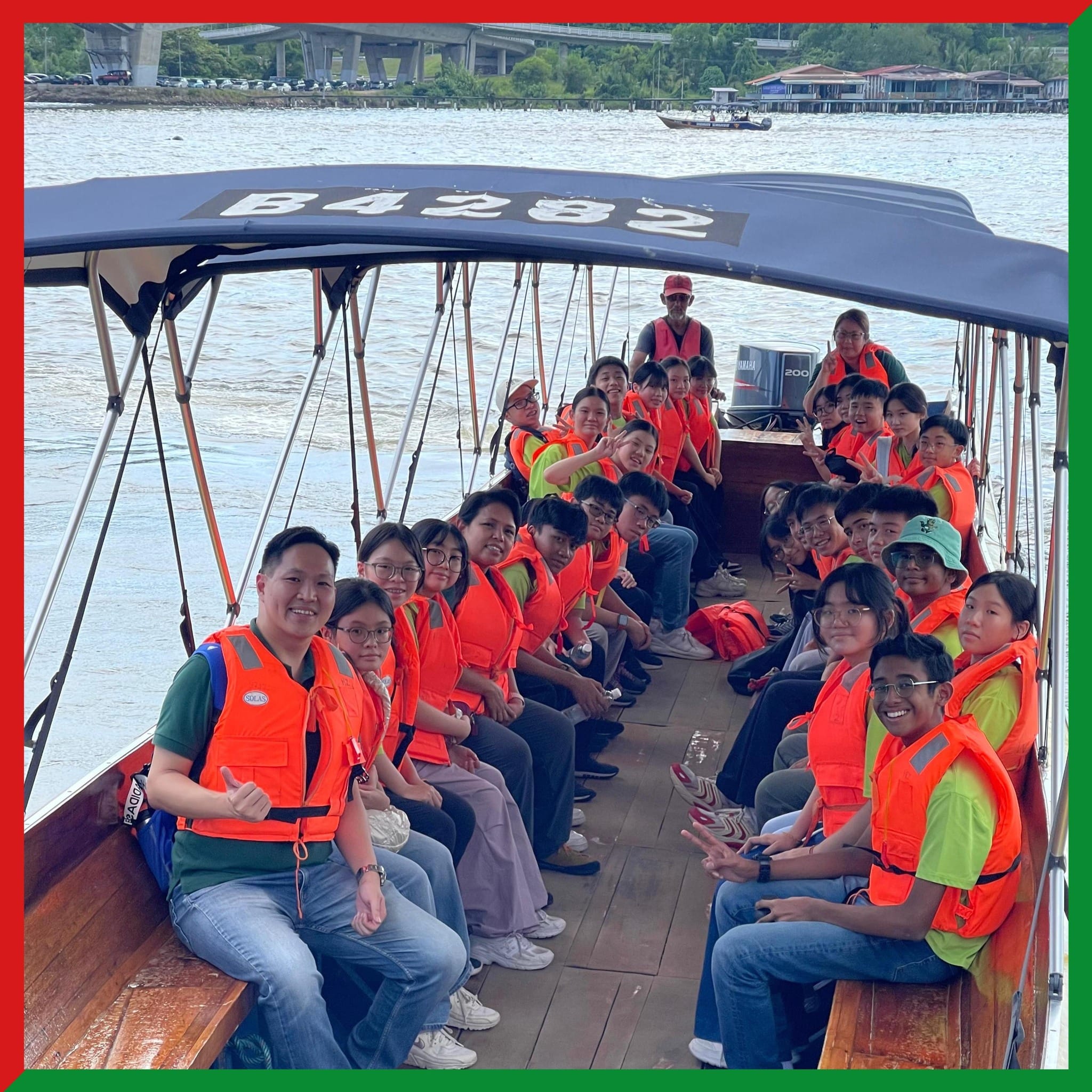 A large group of people wearing orange life jackets seated on a wooden boat labeled B4282 on a river.