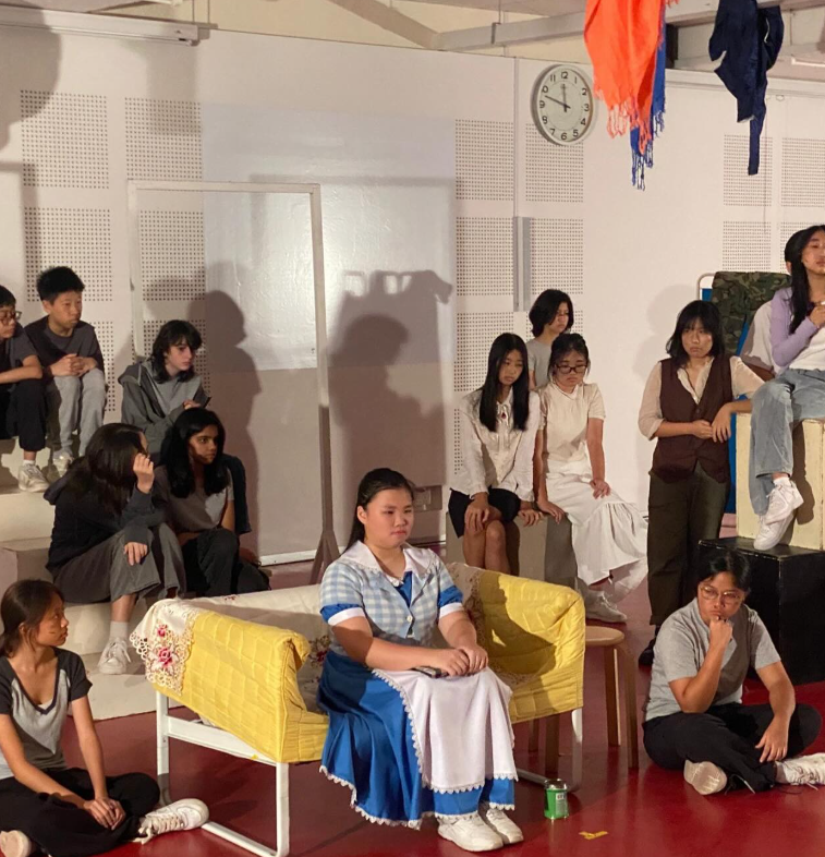 A group of young people on a stage set with a yellow couch, one girl in a blue and white dress seated at center.