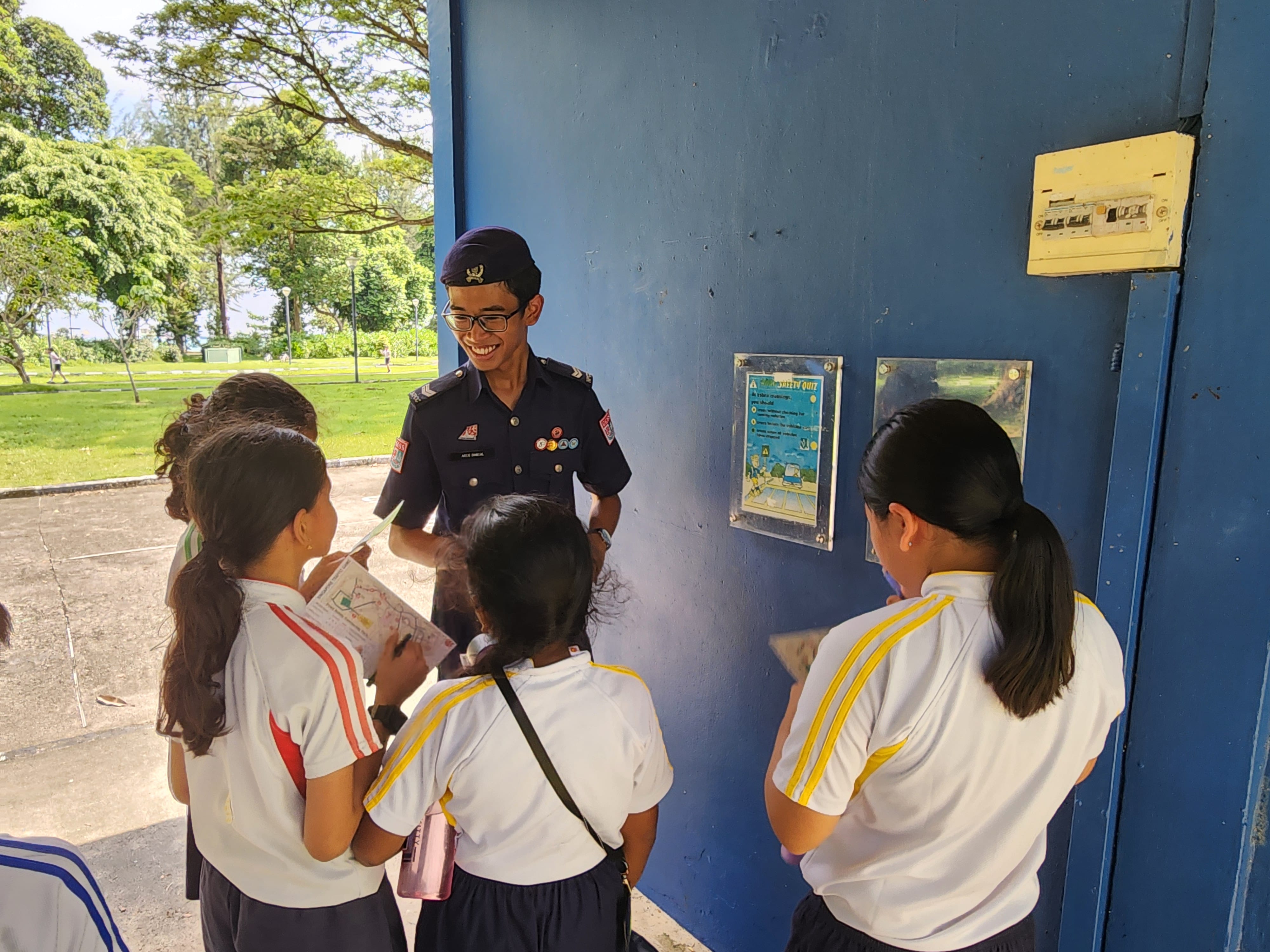 A smiling uniformed cadet with badges talks to a group of young students near a blue wall outdoors.