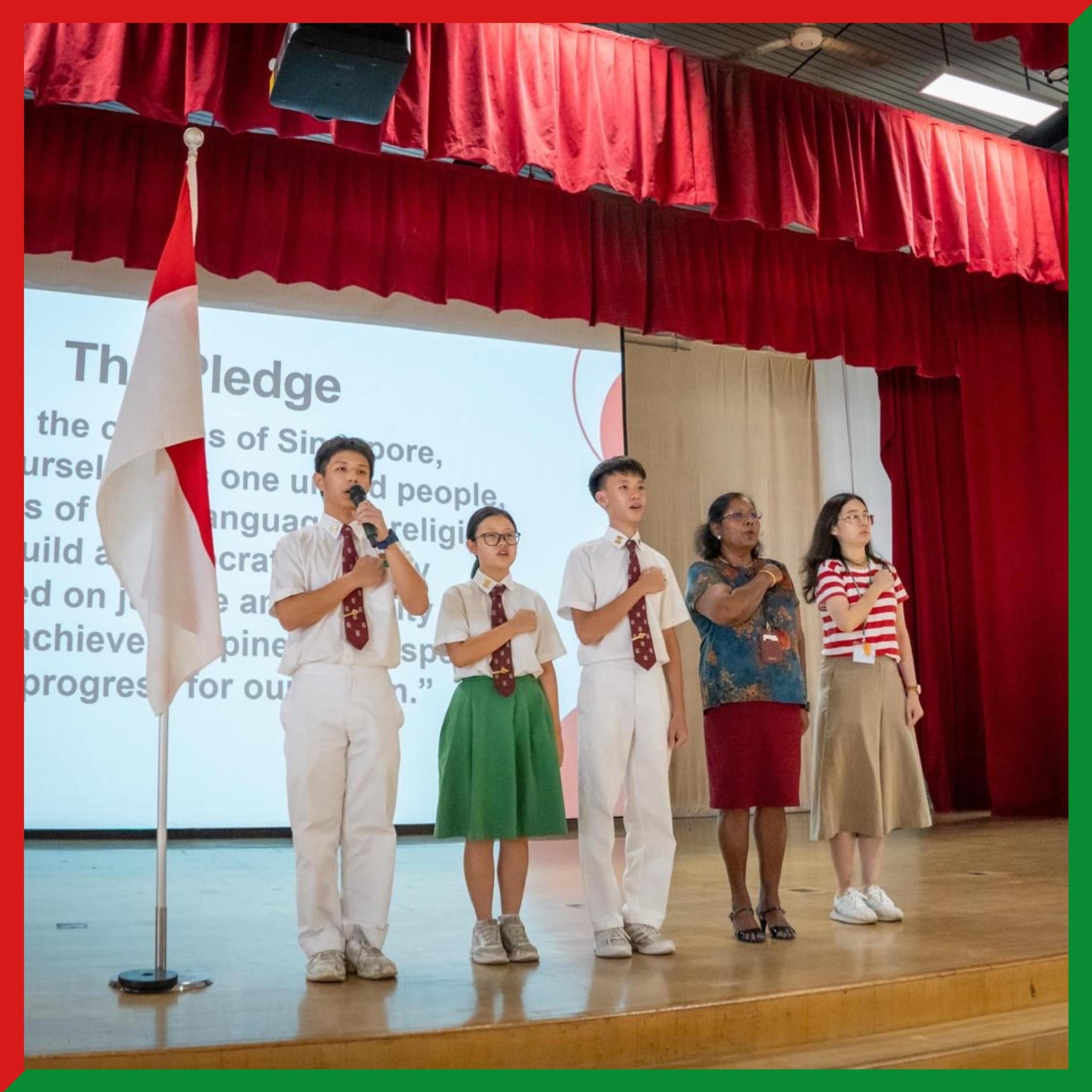 Five people stand on a school stage reciting the Singapore Pledge, with a flag and projected pledge text behind them.