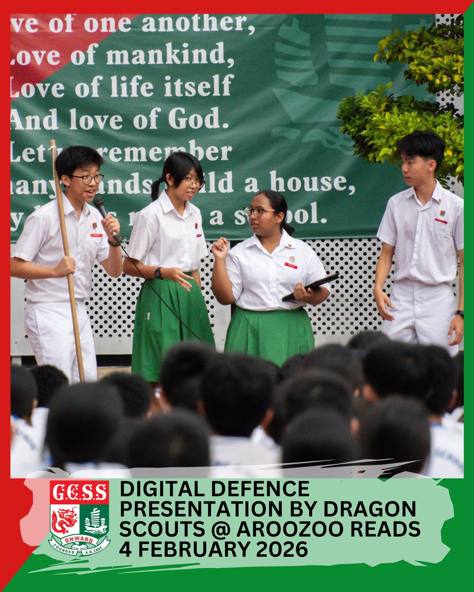 Four students in white and green uniforms present on stage before a seated crowd, with a green banner behind them.