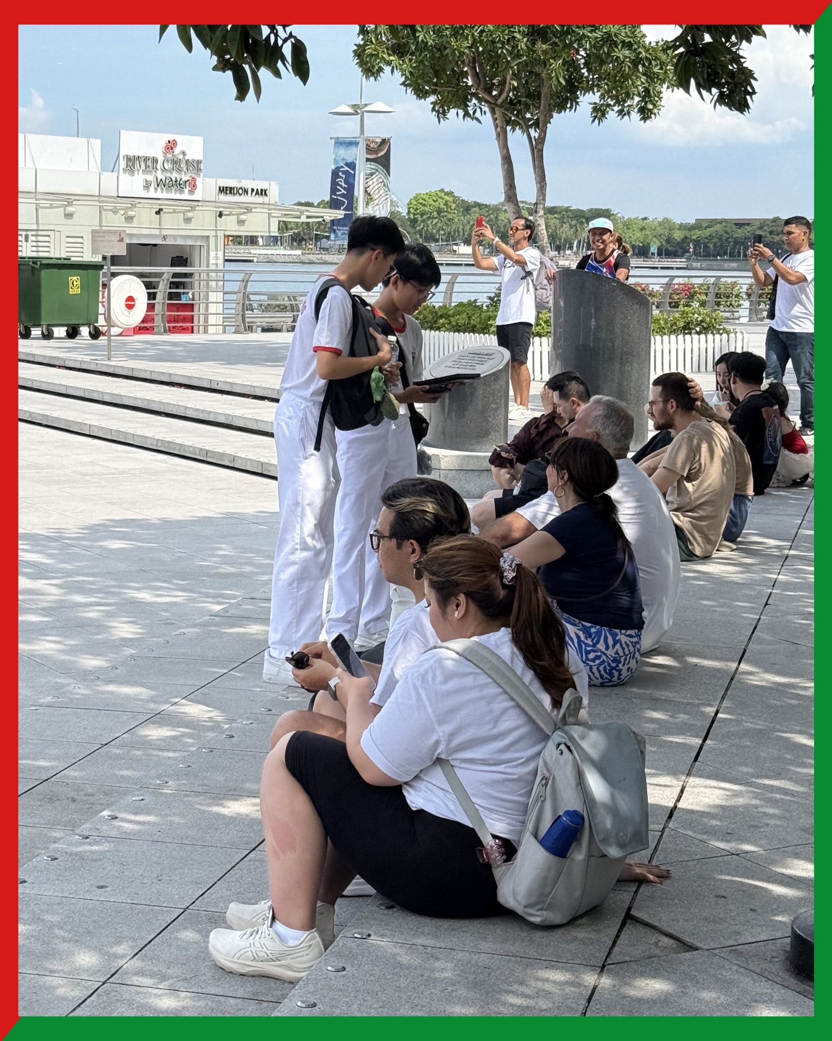 People sitting and standing on a waterfront plaza near Merlion Park in Singapore on a sunny day.