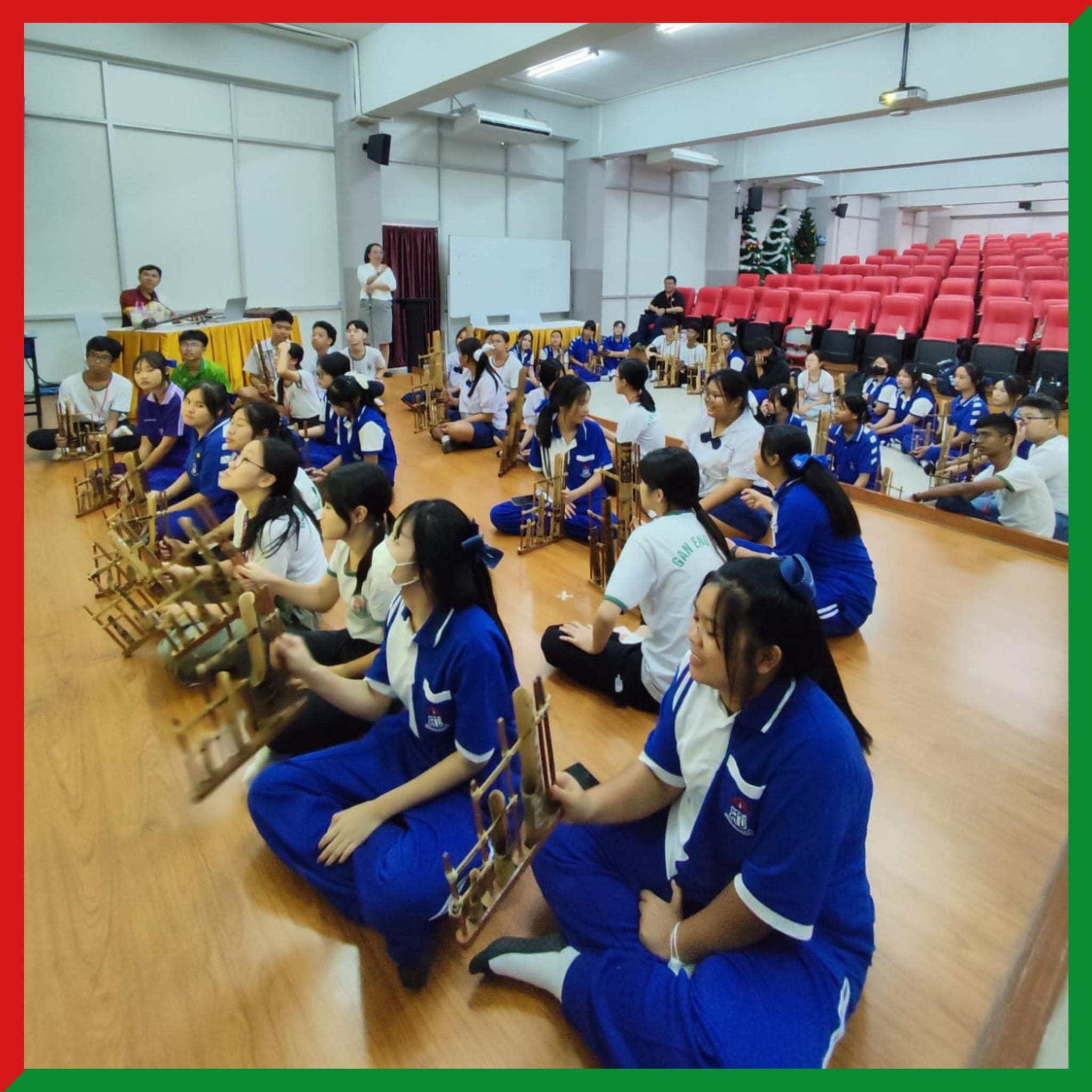 Students in blue uniforms sit on a hall floor holding bamboo instruments during a group music session.