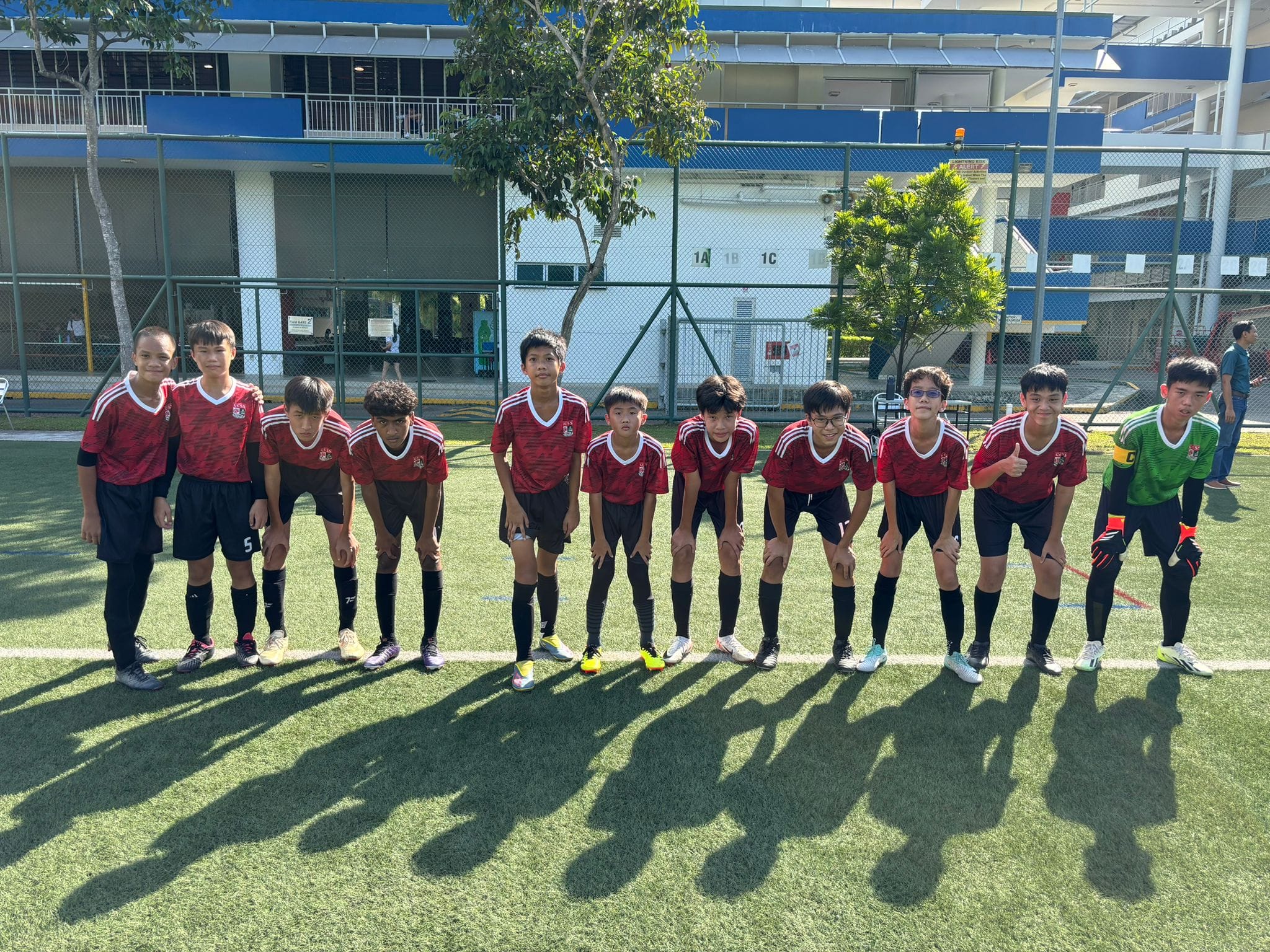 Eleven young boys in red soccer uniforms line up on a green field, with one wearing a green goalkeeper jersey.