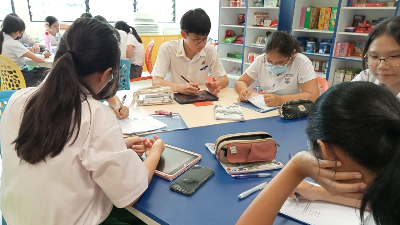 Students in white uniforms sit around a blue table, writing and using tablets in a school canteen.