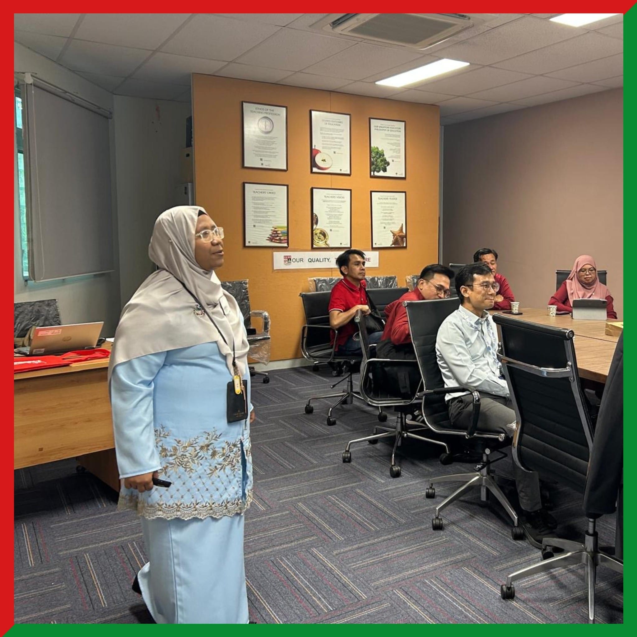 A woman in a blue hijab and dress stands presenting in a meeting room while several people sit at a conference table.