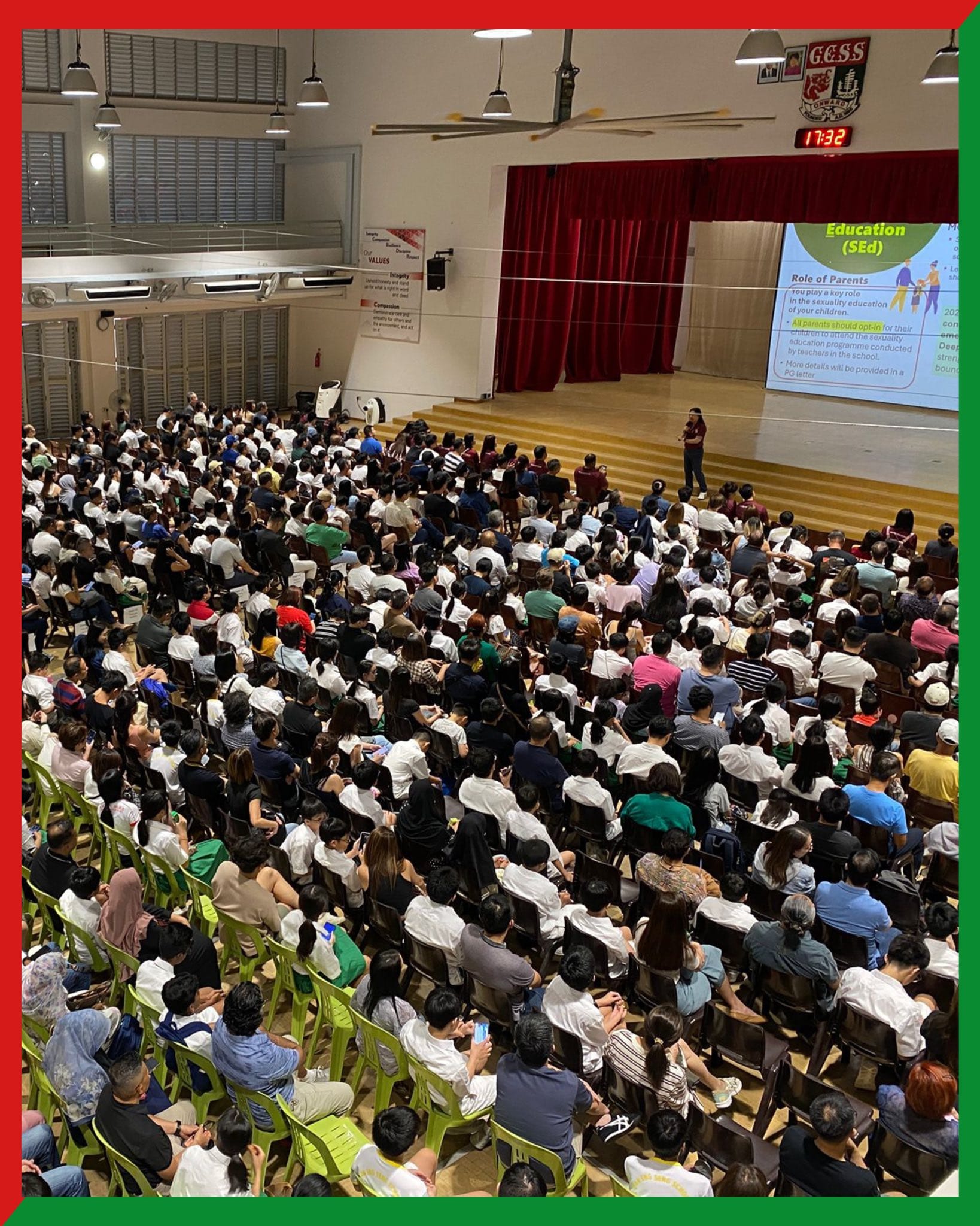 A packed school hall with hundreds of seated attendees facing a presenter on stage with a projected slide about Sexuality Education.