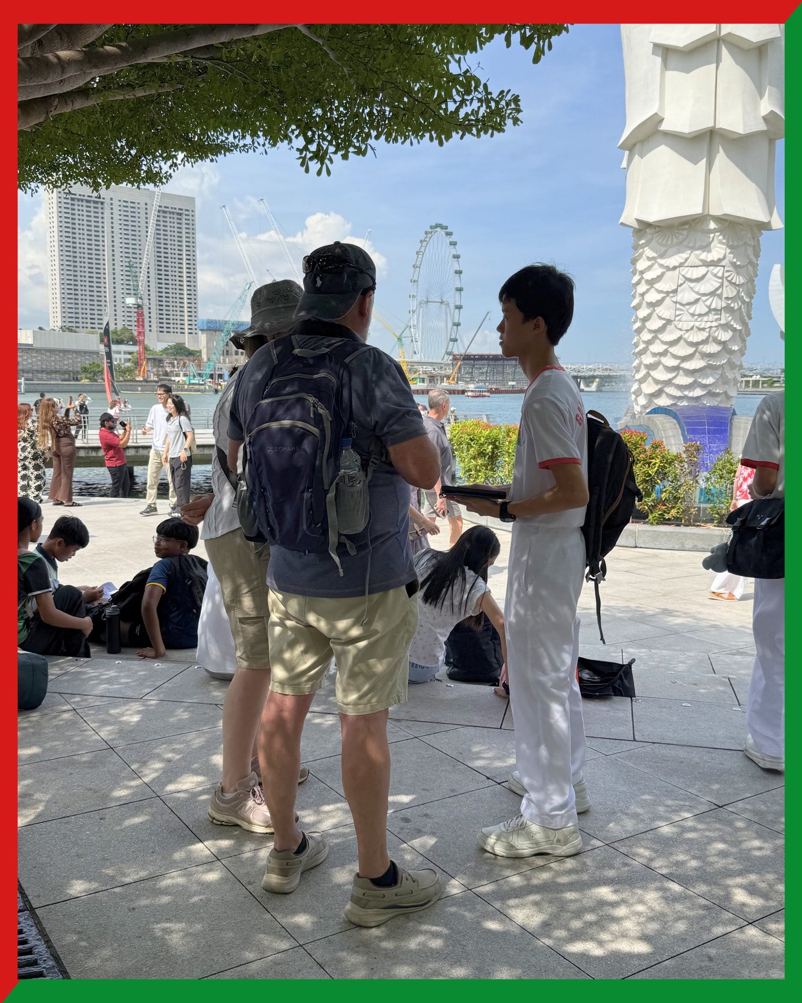 Tourists near the Merlion statue in Singapore with a ferris wheel and waterfront skyline in the background.