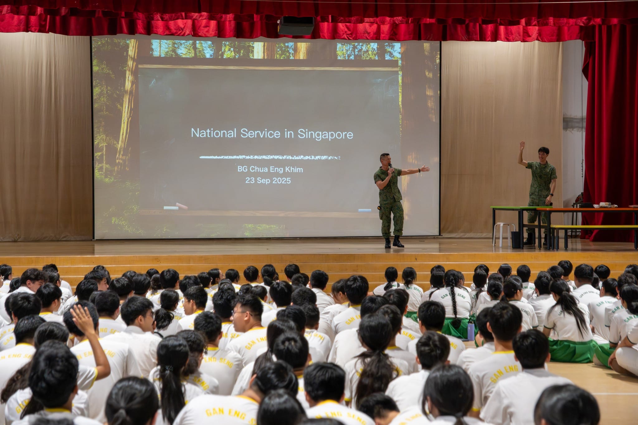 Two uniformed soldiers present a National Service in Singapore talk to students seated on a school hall floor.