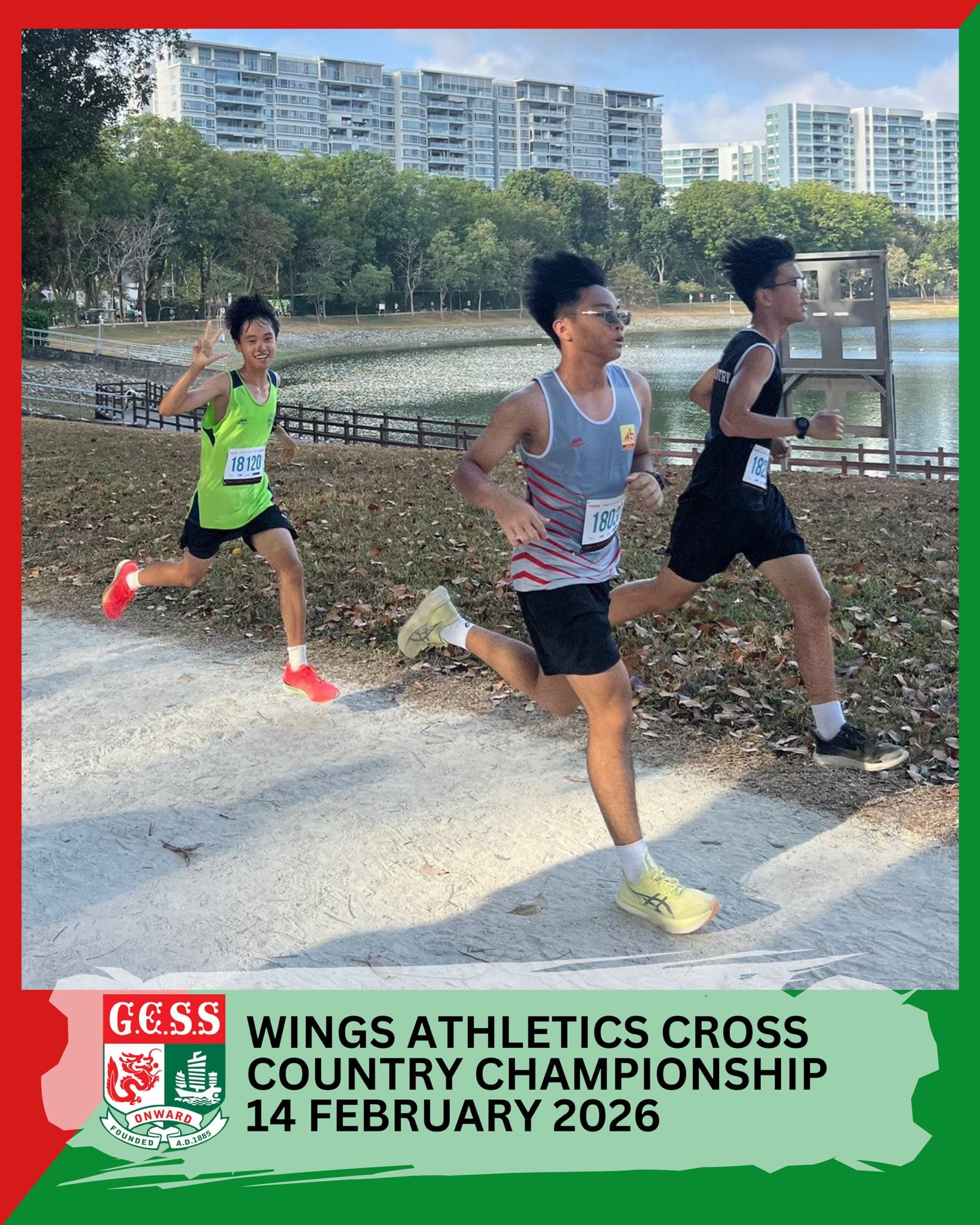 Three young male runners with race bibs compete on a park path near a lake at a cross country championship.