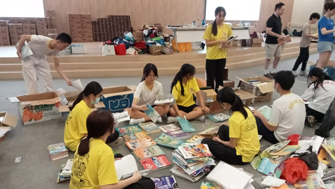 Students in yellow shirts sit on a floor sorting through piles of books and magazines surrounded by cardboard boxes.