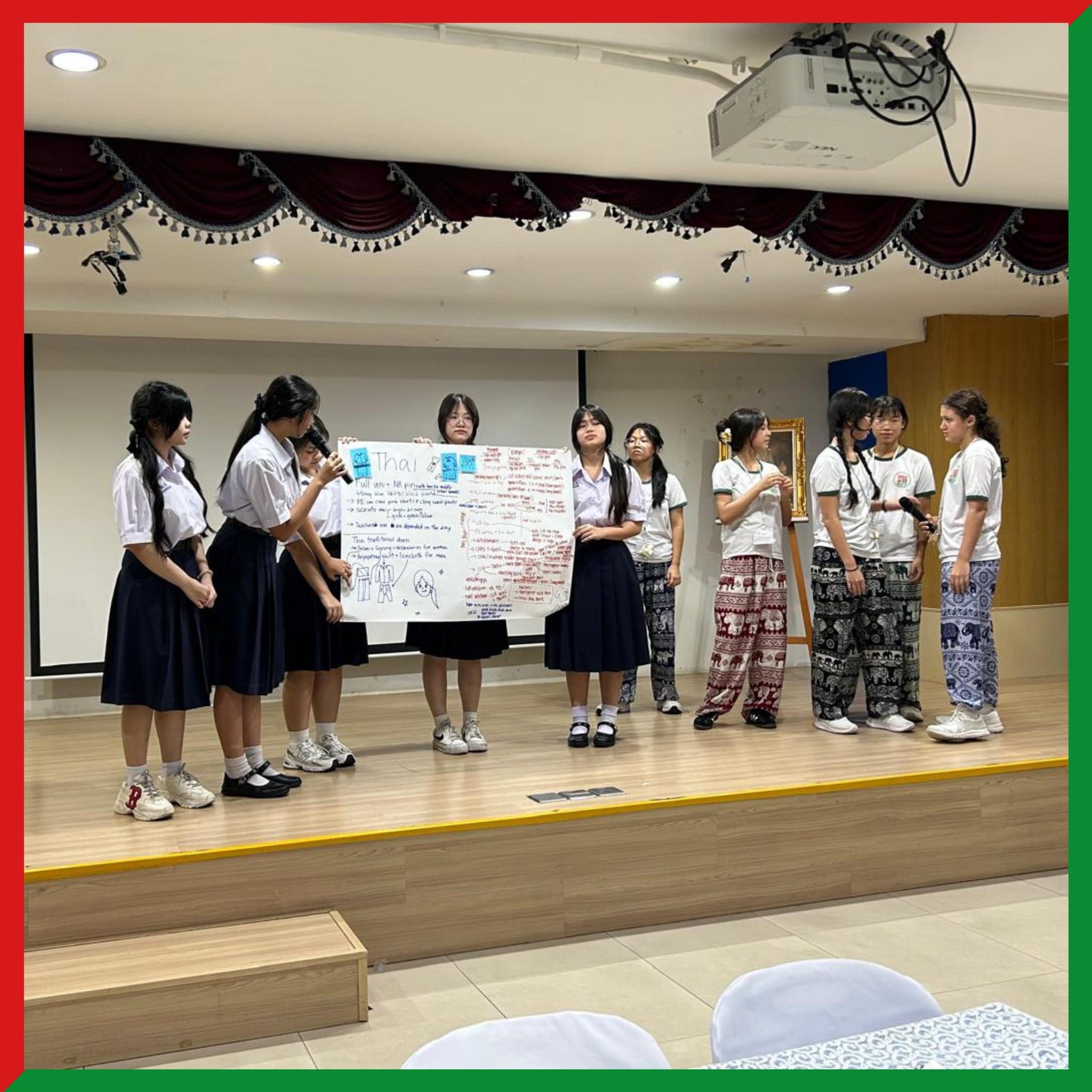 Students in uniforms stand on a stage holding a large handwritten poster labeled Thai during a group presentation.
