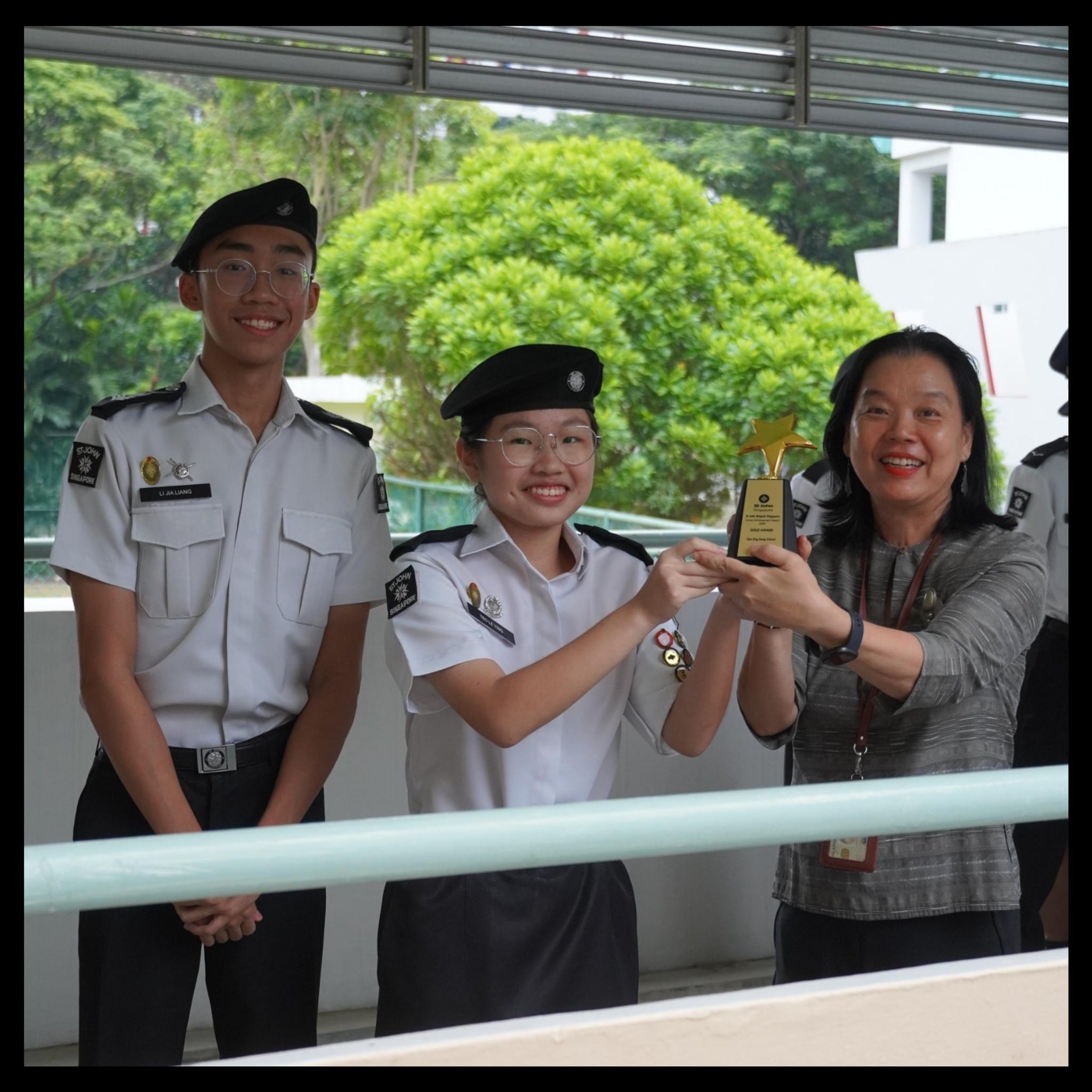 Two uniformed cadets and a smiling woman in grey jointly hold a gold star trophy outdoors.