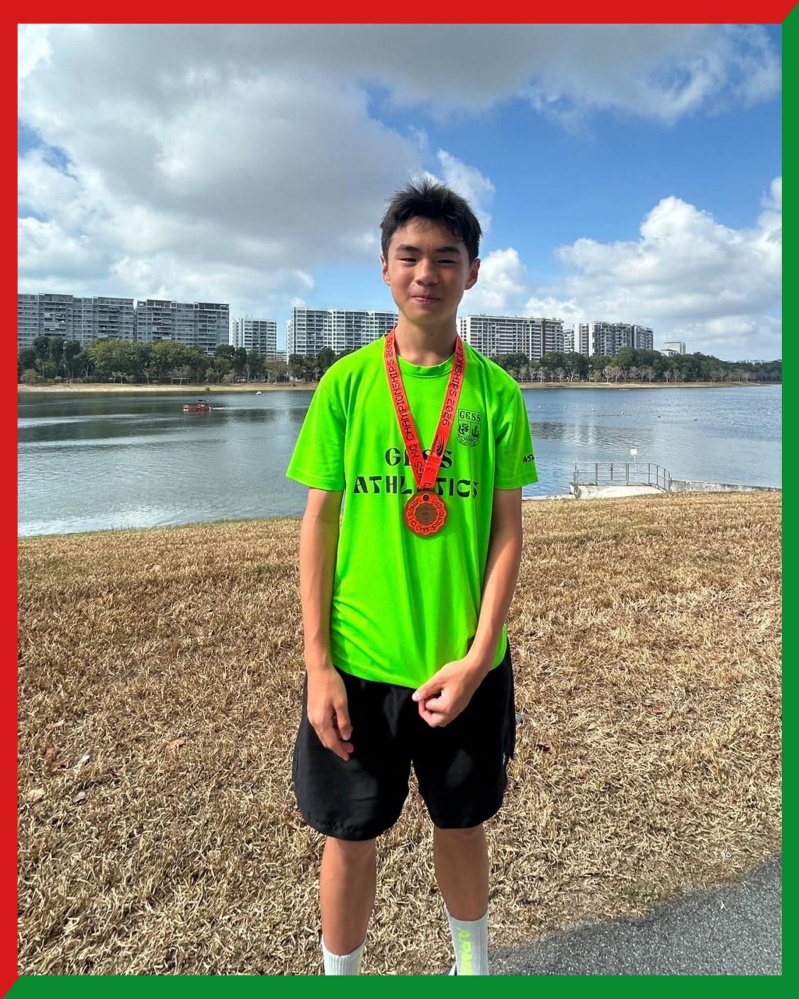 A young male athlete wearing a neon green GESS Athletics shirt and a medal stands by a lake with buildings behind him.
