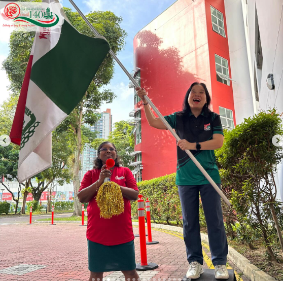 Two women outdoors, one holding a green and white flag on a pole, the other holding a red megaphone with gold pompoms.