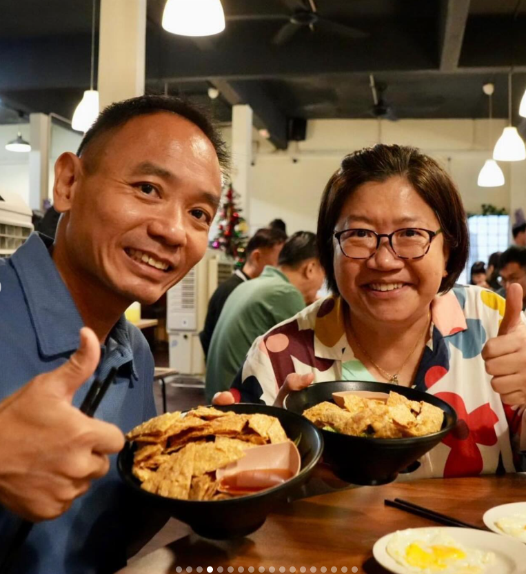 Two smiling people give thumbs up while holding large bowls of food at a restaurant table.