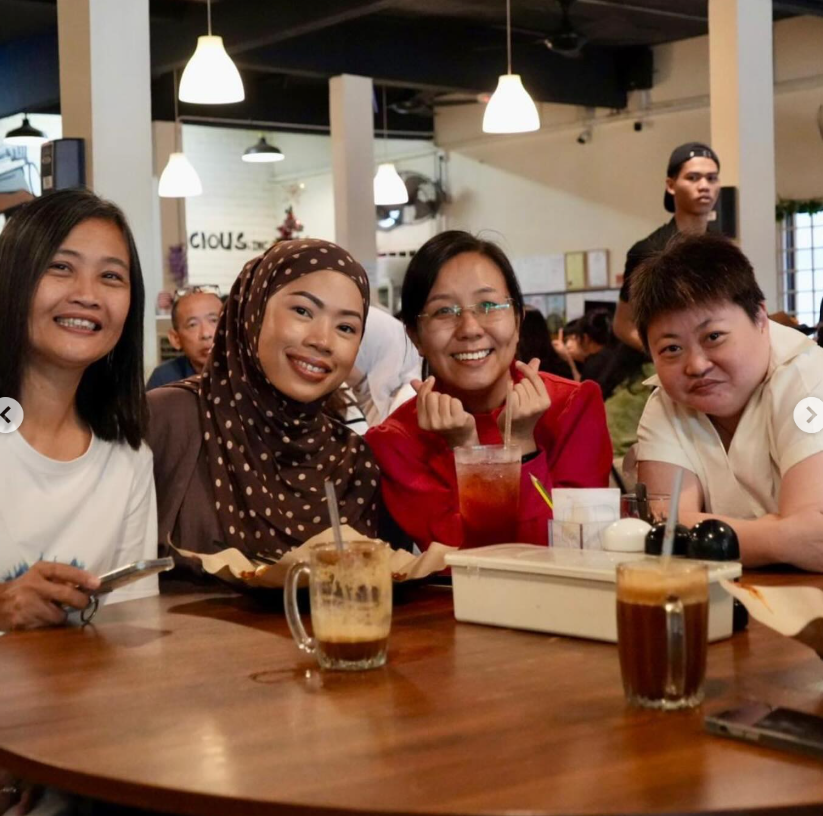 Four smiling women sit together at a wooden table in a cafe, with drinks in front of them.