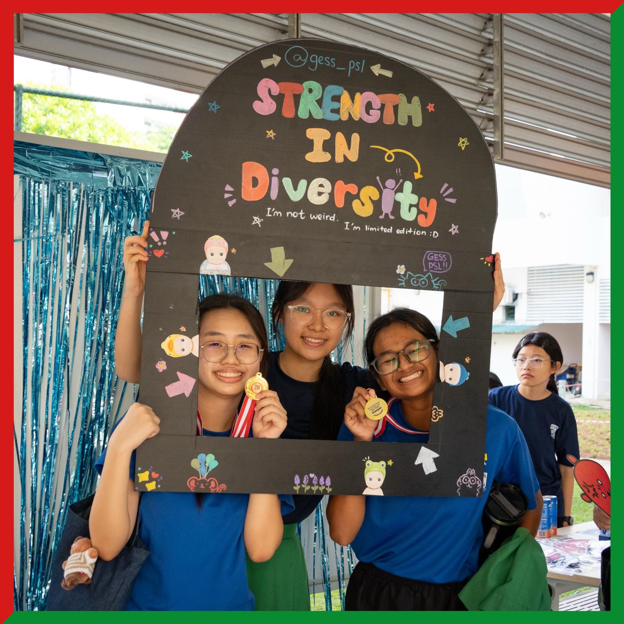 Three smiling students hold medals inside a decorated cardboard frame reading "Strength in Diversity" at a school event.