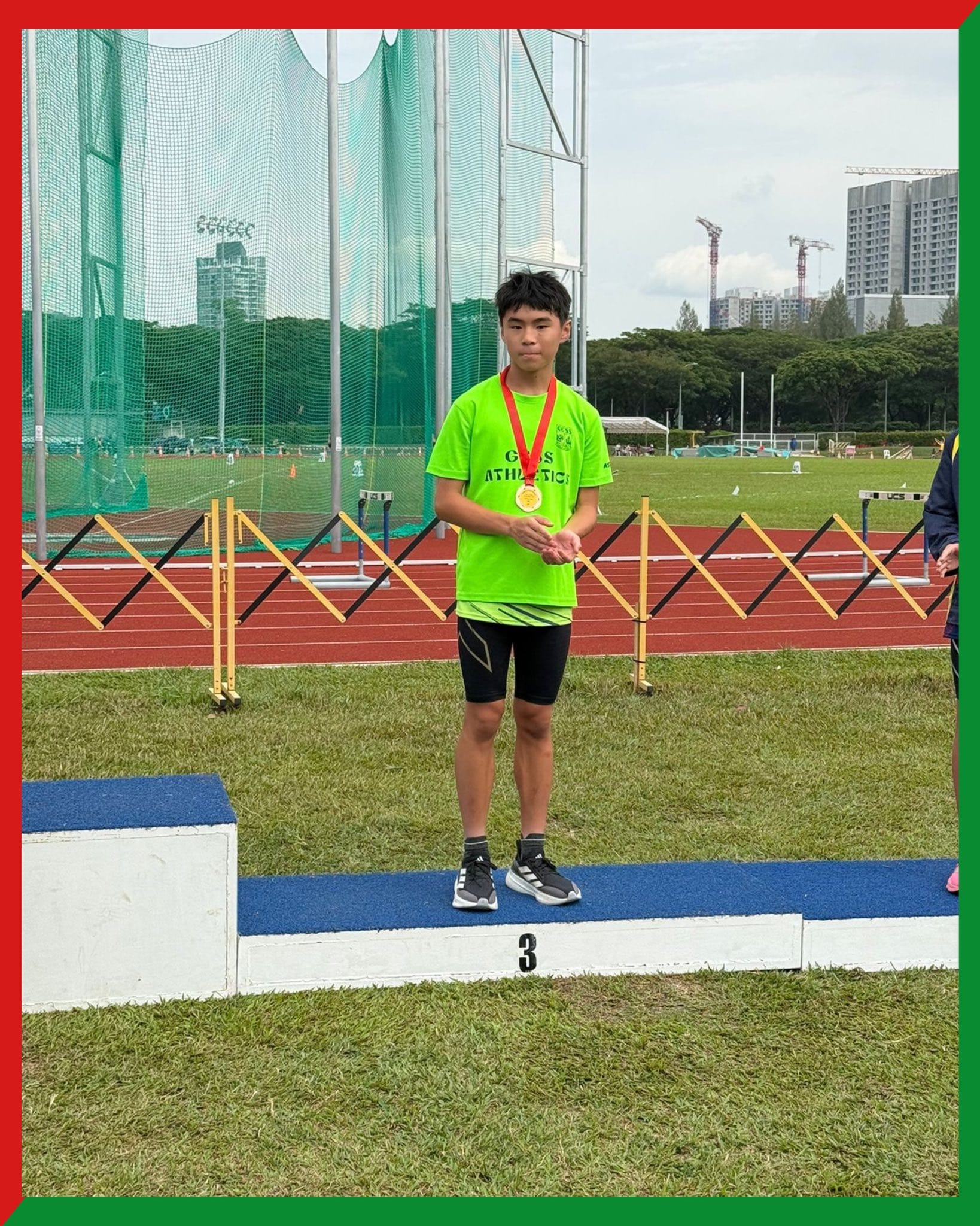 A young male athlete in a green jersey stands on the third-place podium wearing a gold medal at a track meet.