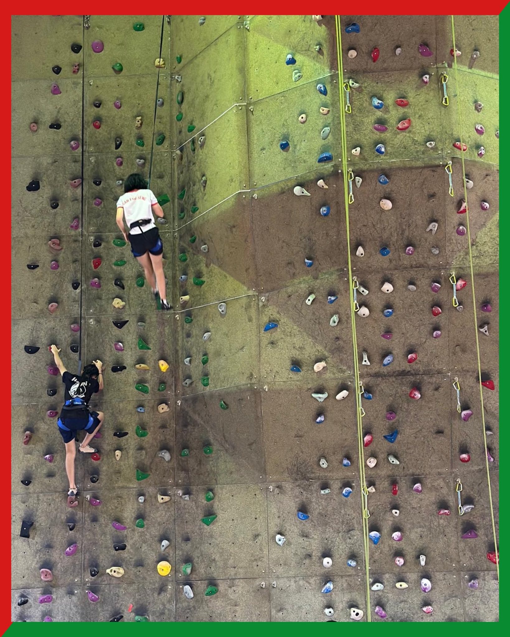 Two people climbing an indoor rock wall with colorful holds, one higher up in white shirt, one lower in black shirt.