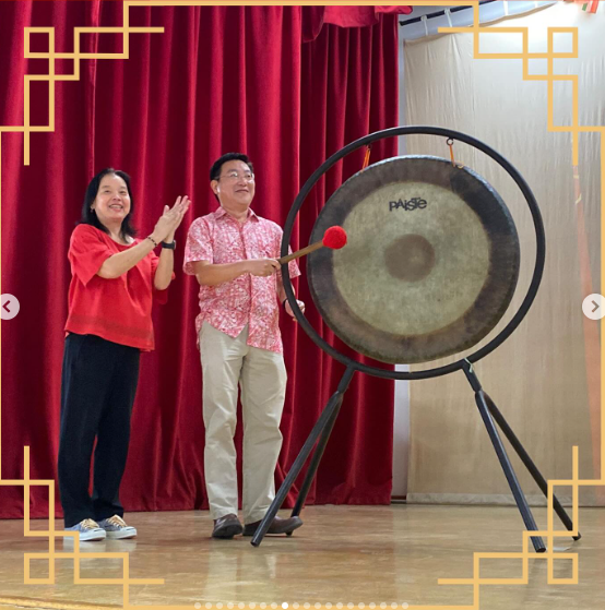 Two people smile near a large Paiste gong on a stand, with a red velvet curtain behind them.