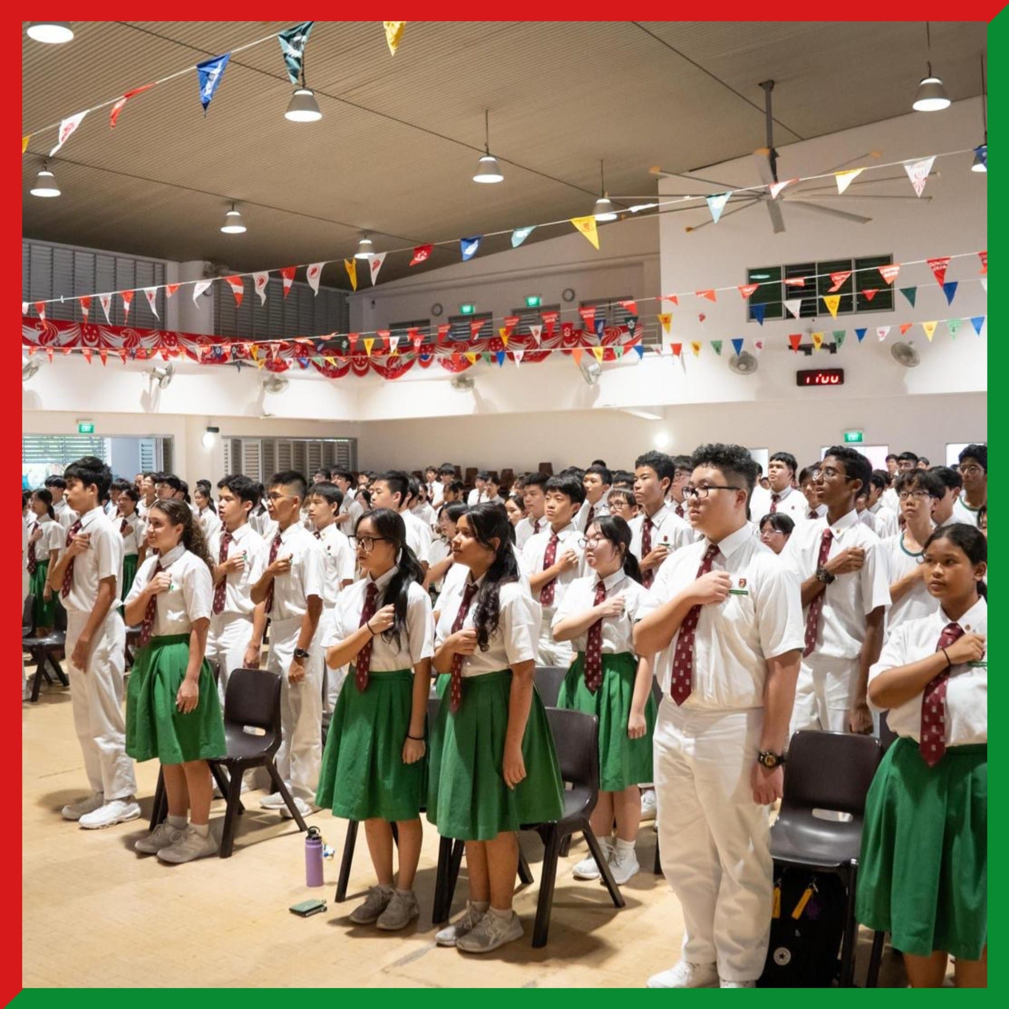 Students in white shirts, red ties, and green skirts stand with hands on chests in a decorated school hall.