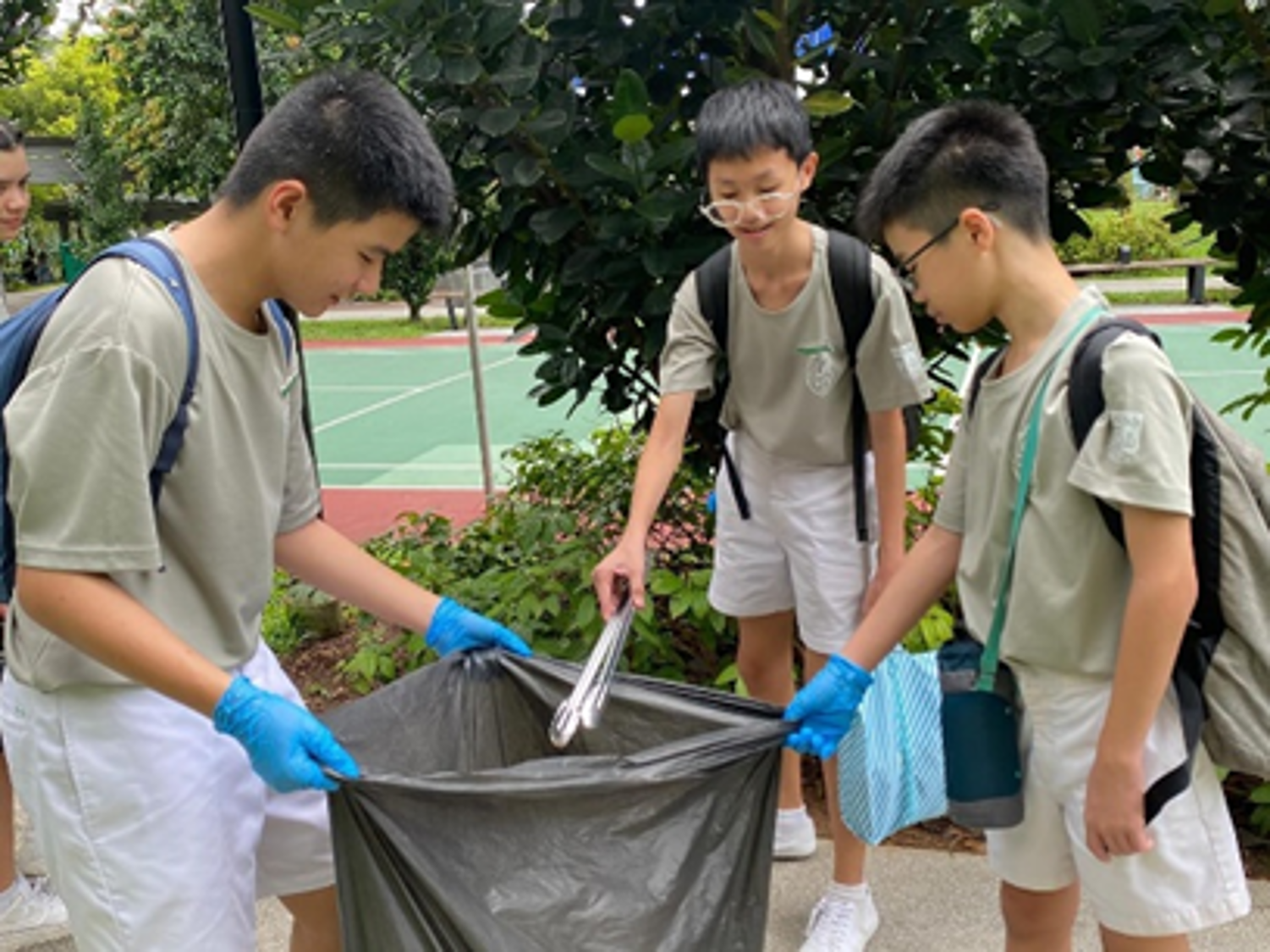 Three boys in school uniforms and blue gloves hold a trash bag as one uses tongs to pick up litter.