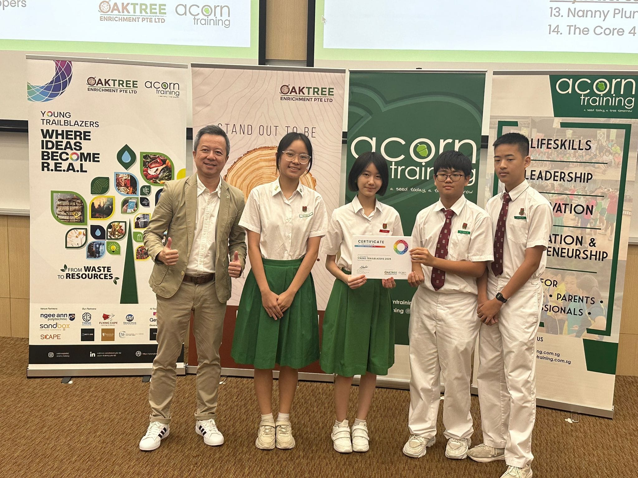 An adult man and four students in white uniforms pose with a certificate in front of Oaktree and Acorn Training banners.