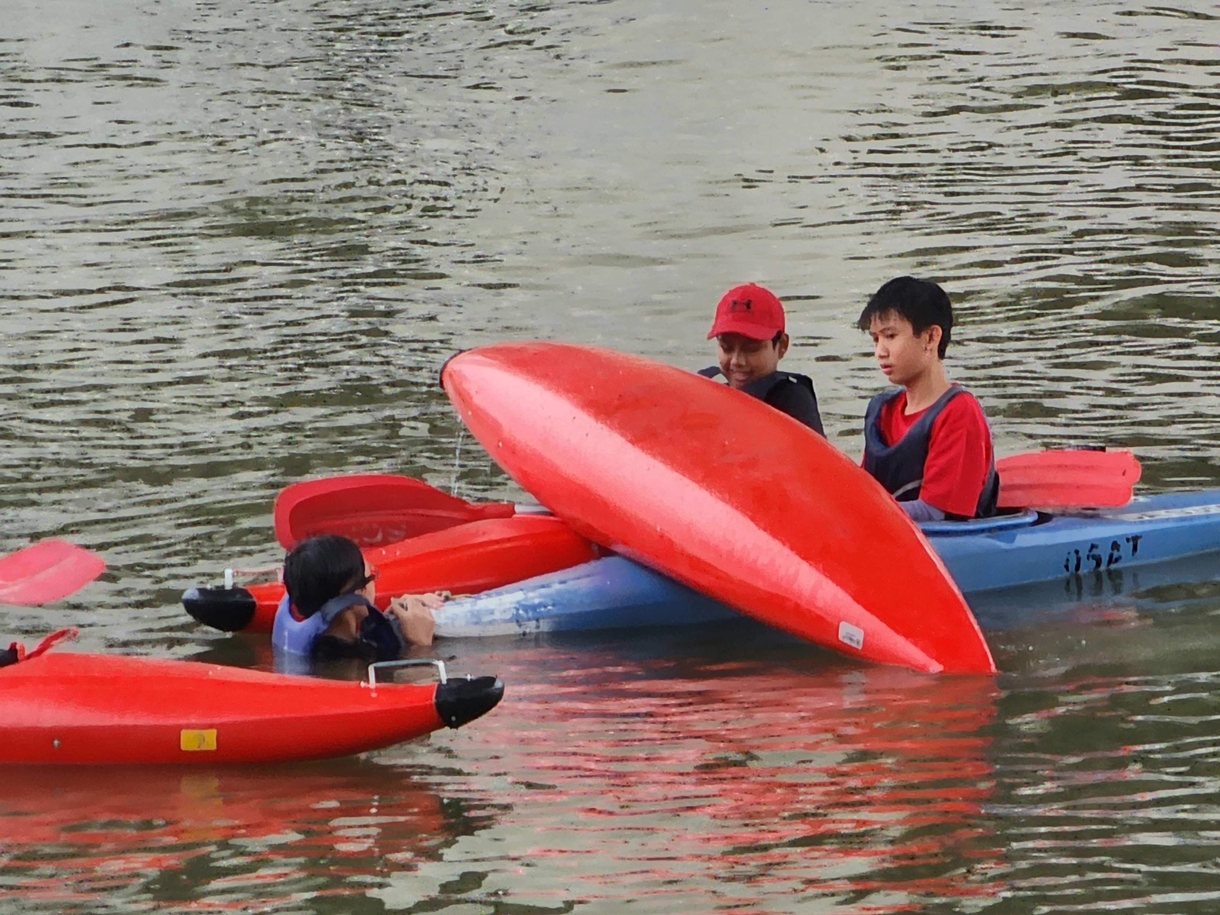 Three children in life vests on water with red and blue kayaks, one kayak flipped upside down.