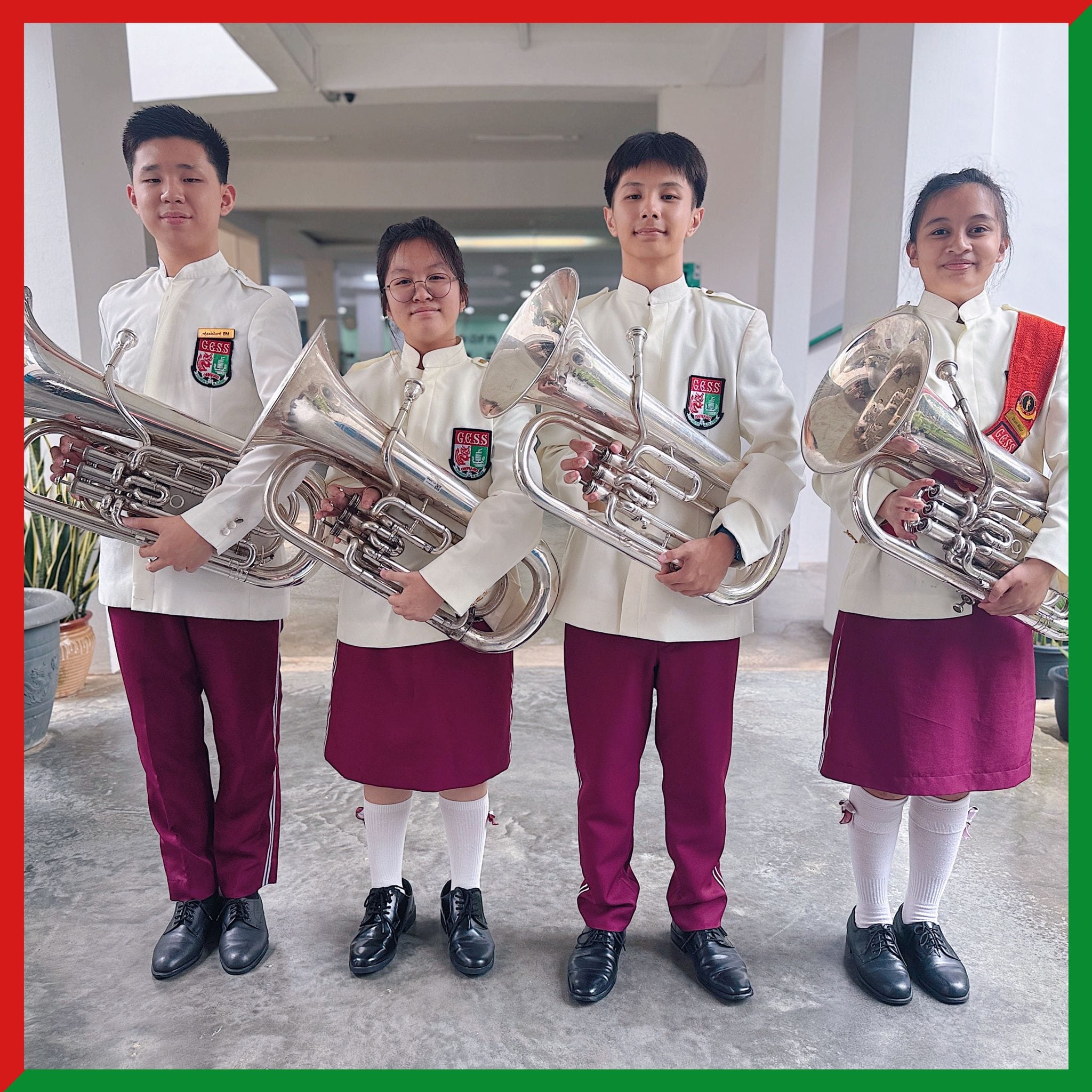 Four students in white and maroon school band uniforms holding large brass instruments indoors.