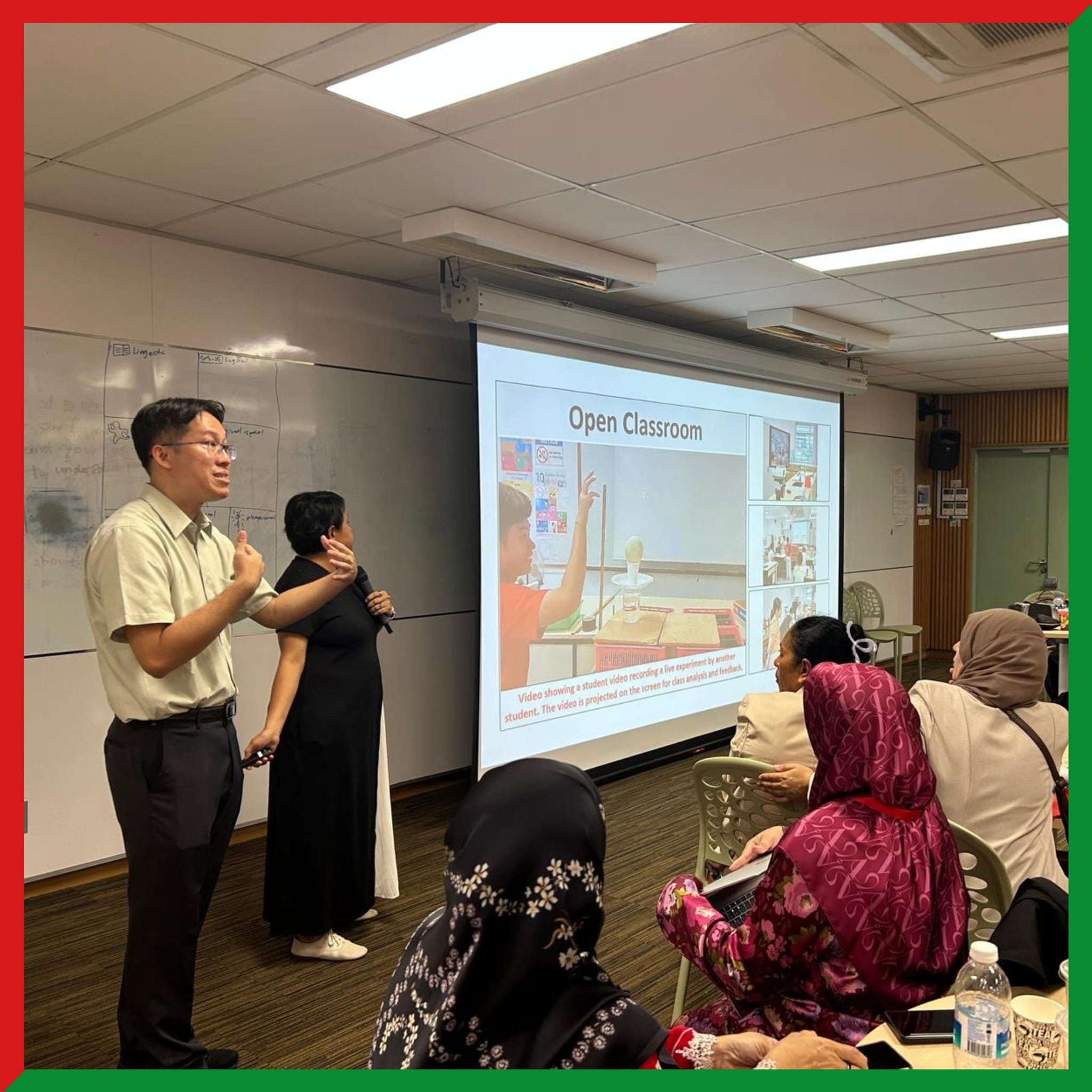 Two presenters stand before a projected slide titled Open Classroom while attendees sit and listen in a meeting room.