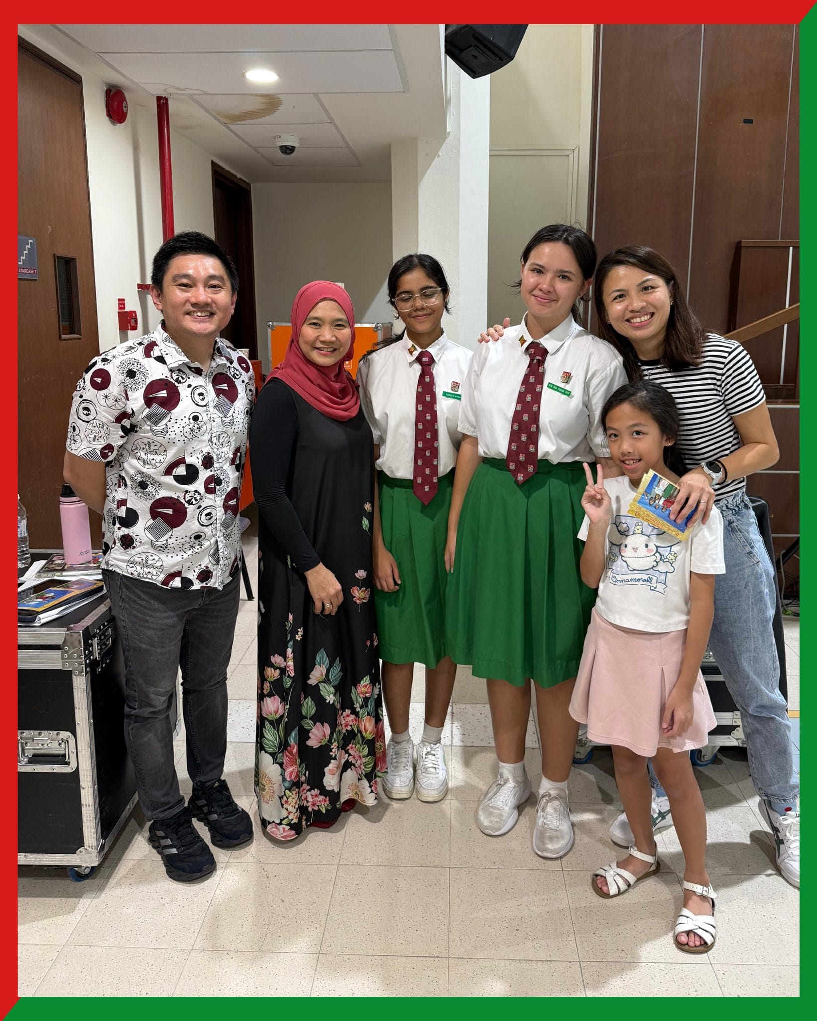 Six people pose together indoors, including two students in white shirts, green skirts, and red ties, and a young girl making a peace sign.