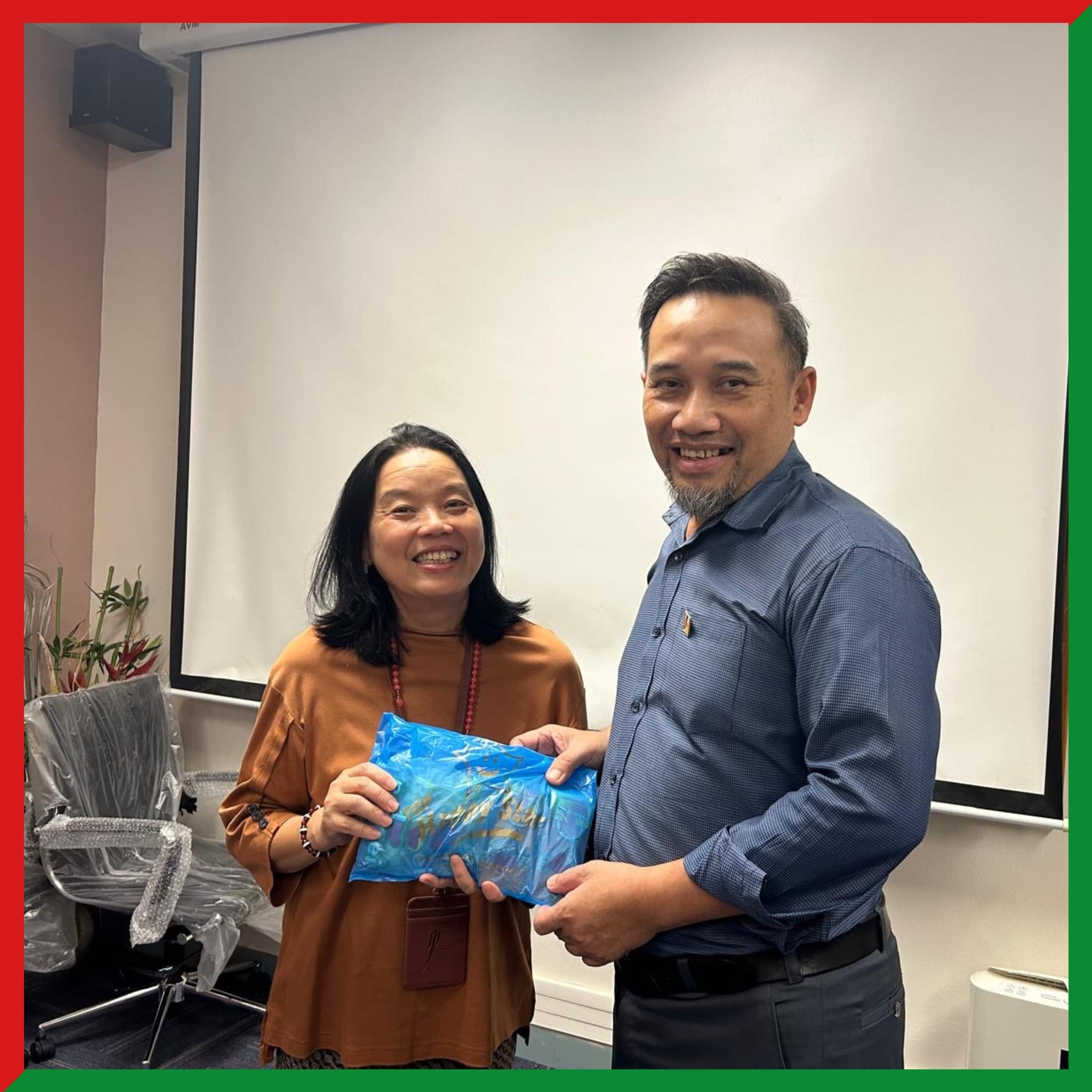 Two smiling people stand together in a conference room, jointly holding a blue gift bag.