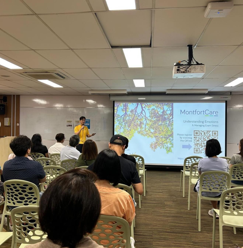 A speaker in a yellow shirt presents a MontfortCare slide on managing exam stress to a seated audience in a meeting room.
