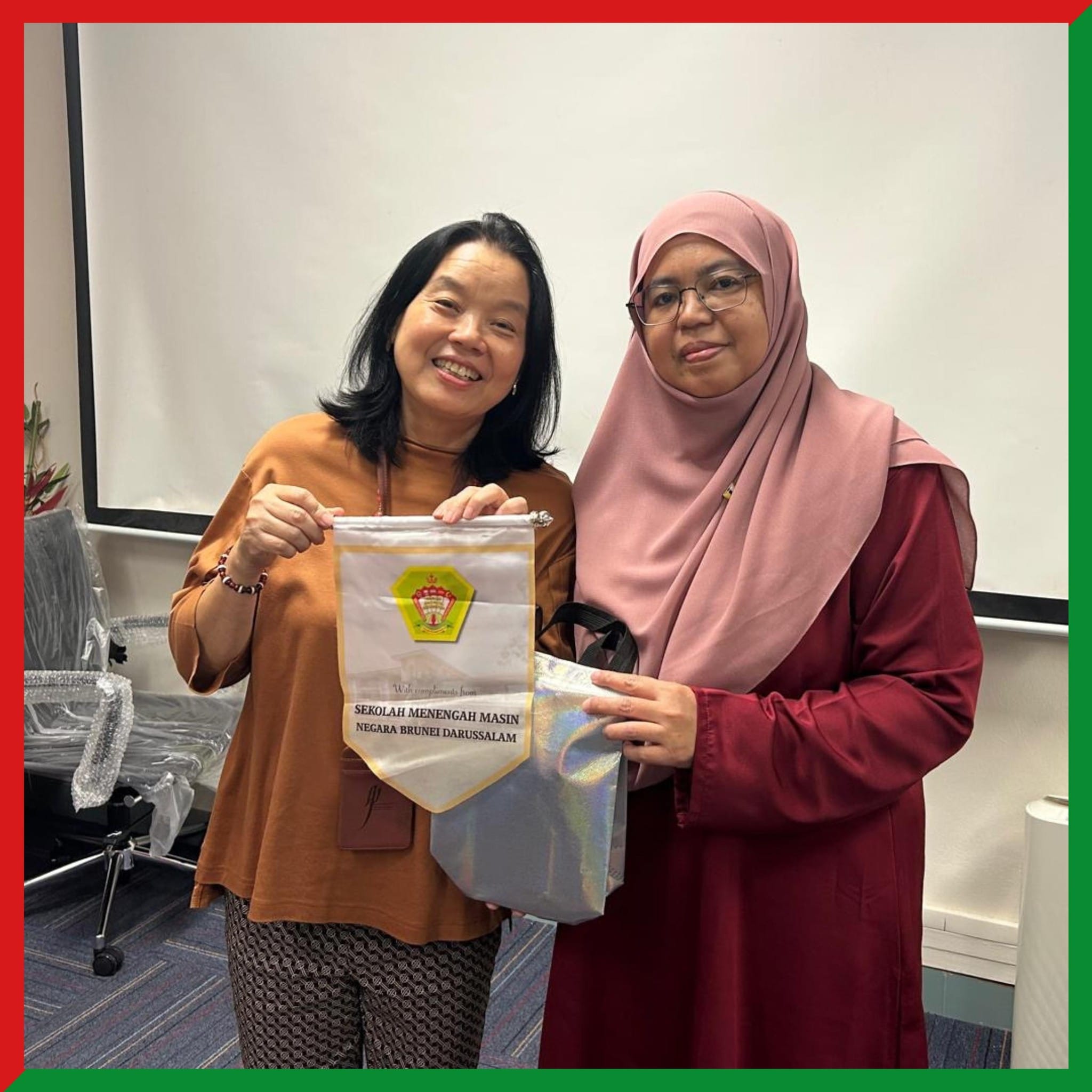 Two women pose together holding a pennant reading Sekolah Menengah Masin, Negara Brunei Darussalam.