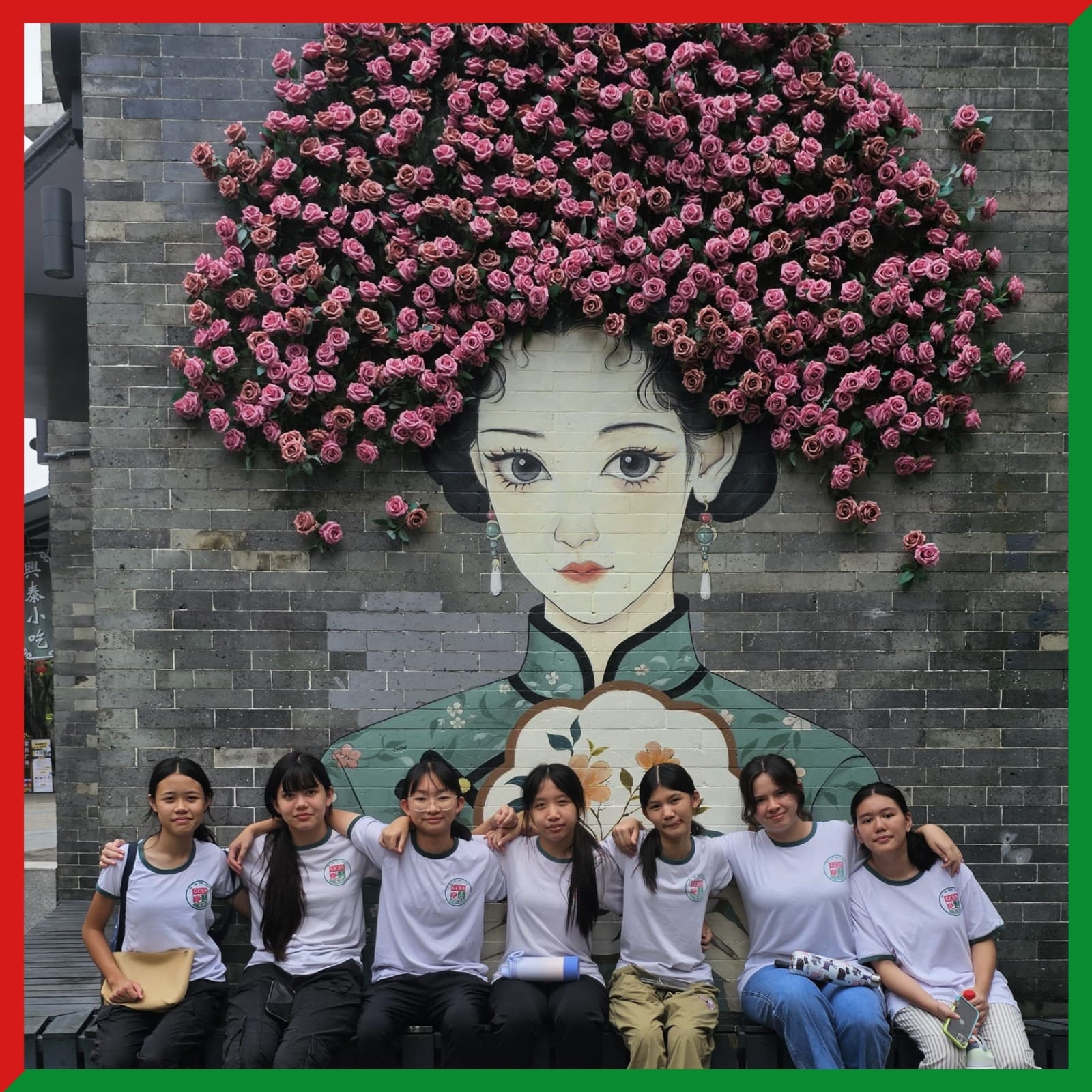 Seven young women in matching white shirts sit in front of a brick wall mural of a woman with pink roses as hair.