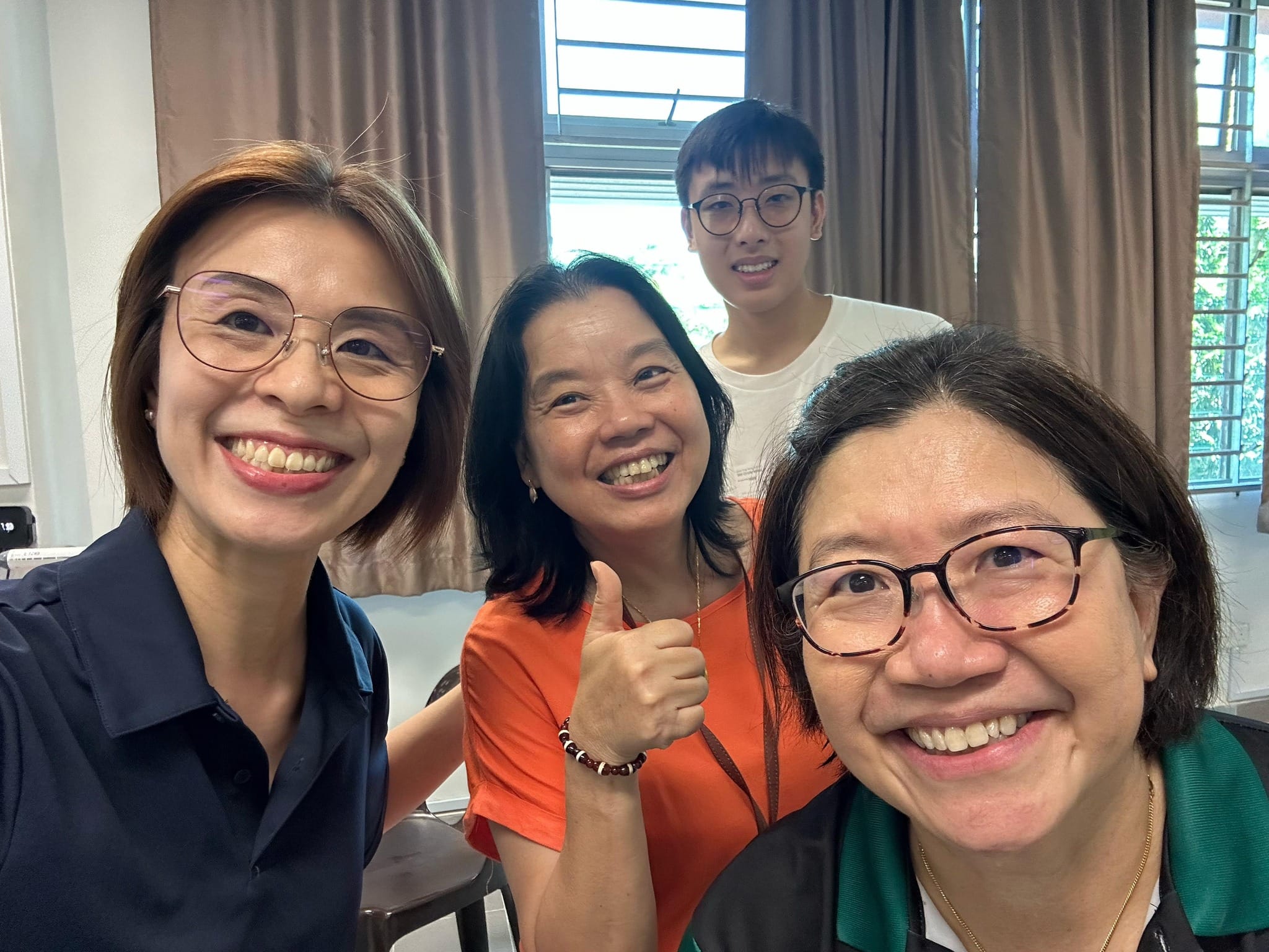 Four smiling people pose for a selfie indoors, with one woman in orange giving a thumbs up.