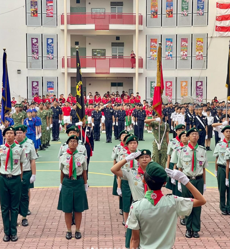 Students in scout and military uniforms stand at attention and salute during a school parade ceremony outdoors.