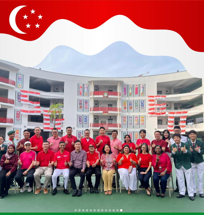 Group of people in red attire posing with thumbs up in front of a school decorated with Singapore flags.