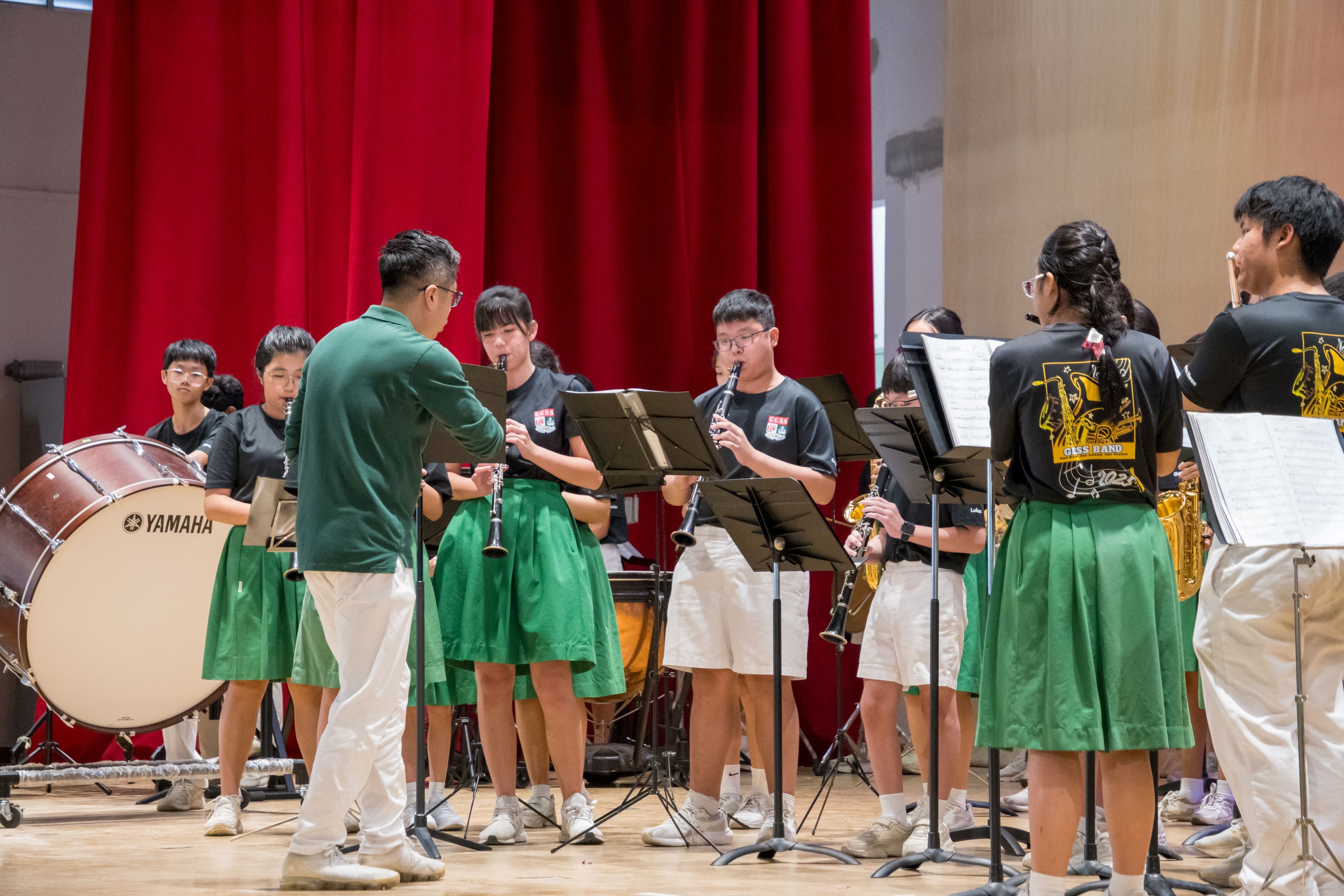 A youth band in green and black uniforms plays clarinets and percussion on a stage with a red curtain backdrop.