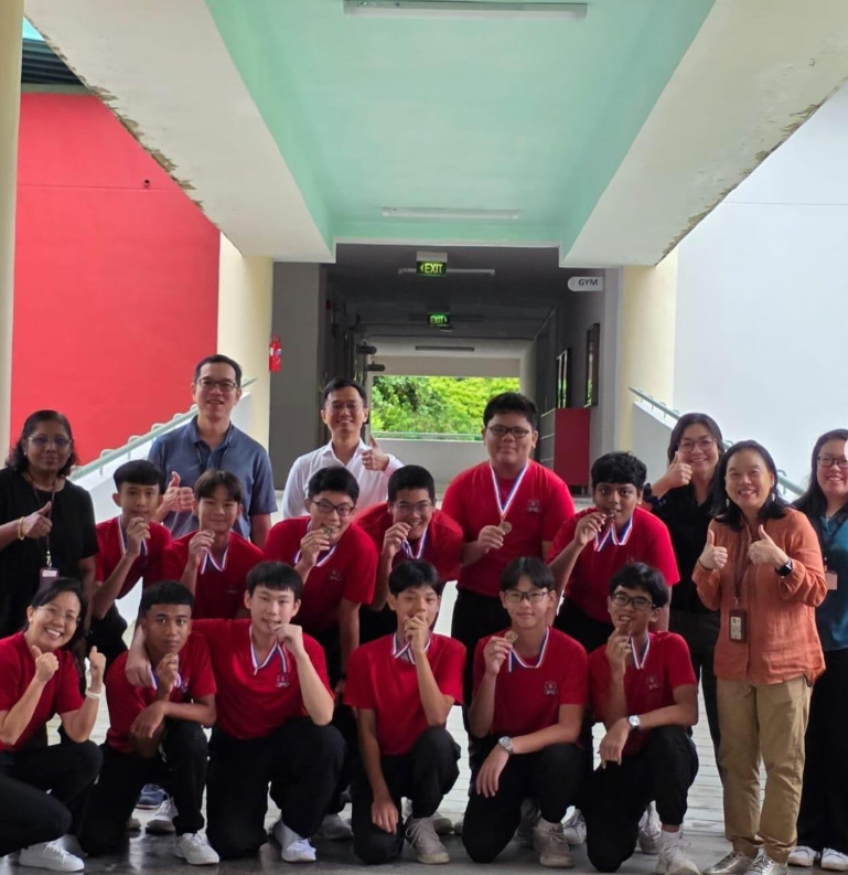 A group of students in red uniforms holding medals pose with thumbs up alongside several adults in a school corridor.