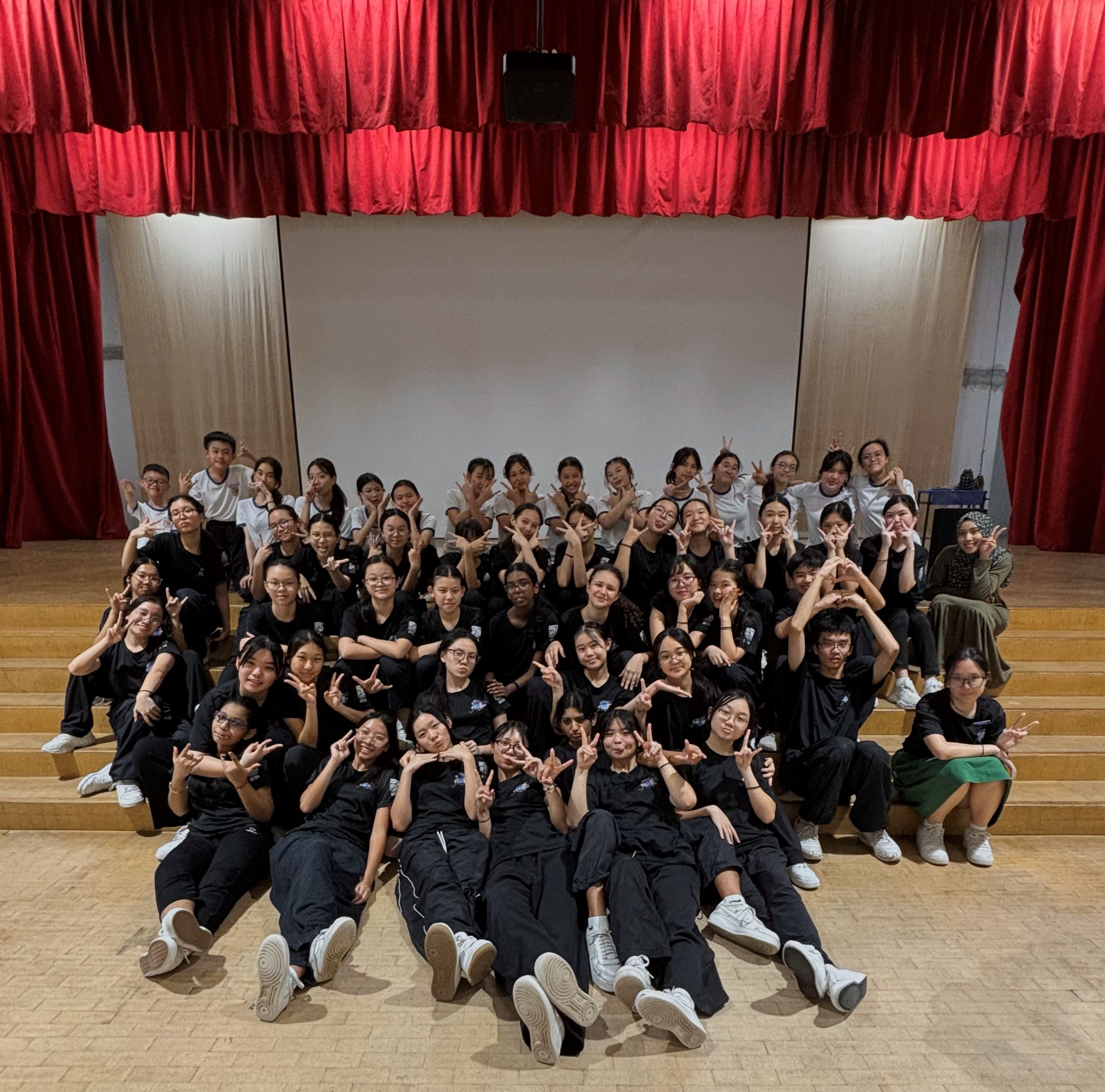 Large group of young people in black and white outfits posing on a stage with red curtains, making hand gestures.