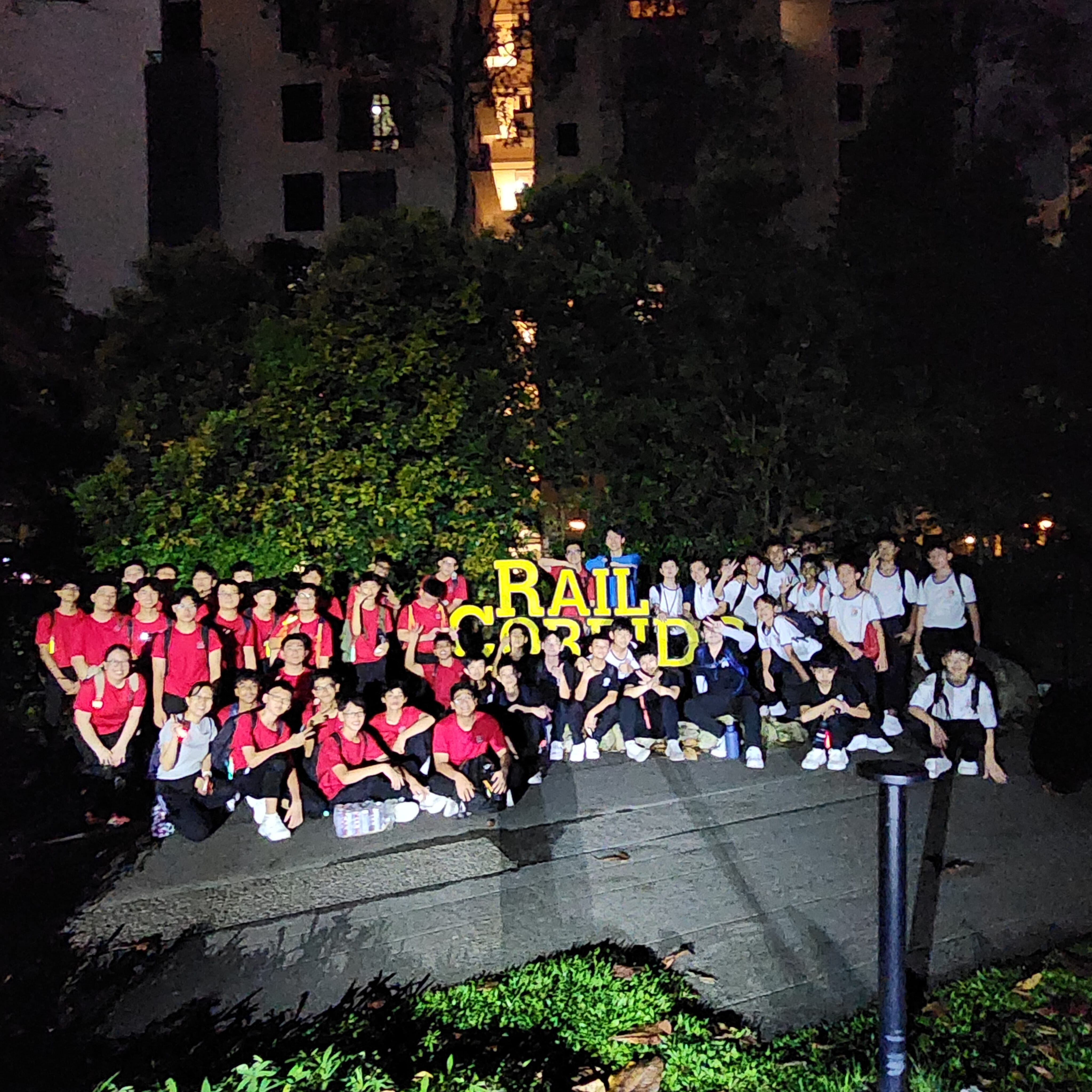 Large group of young people in red and white shirts posing at night by a yellow Rail Corridor sign amid trees.