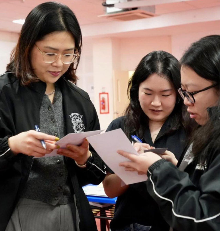 Three young women in black jackets look at papers together, two holding pens, standing indoors.