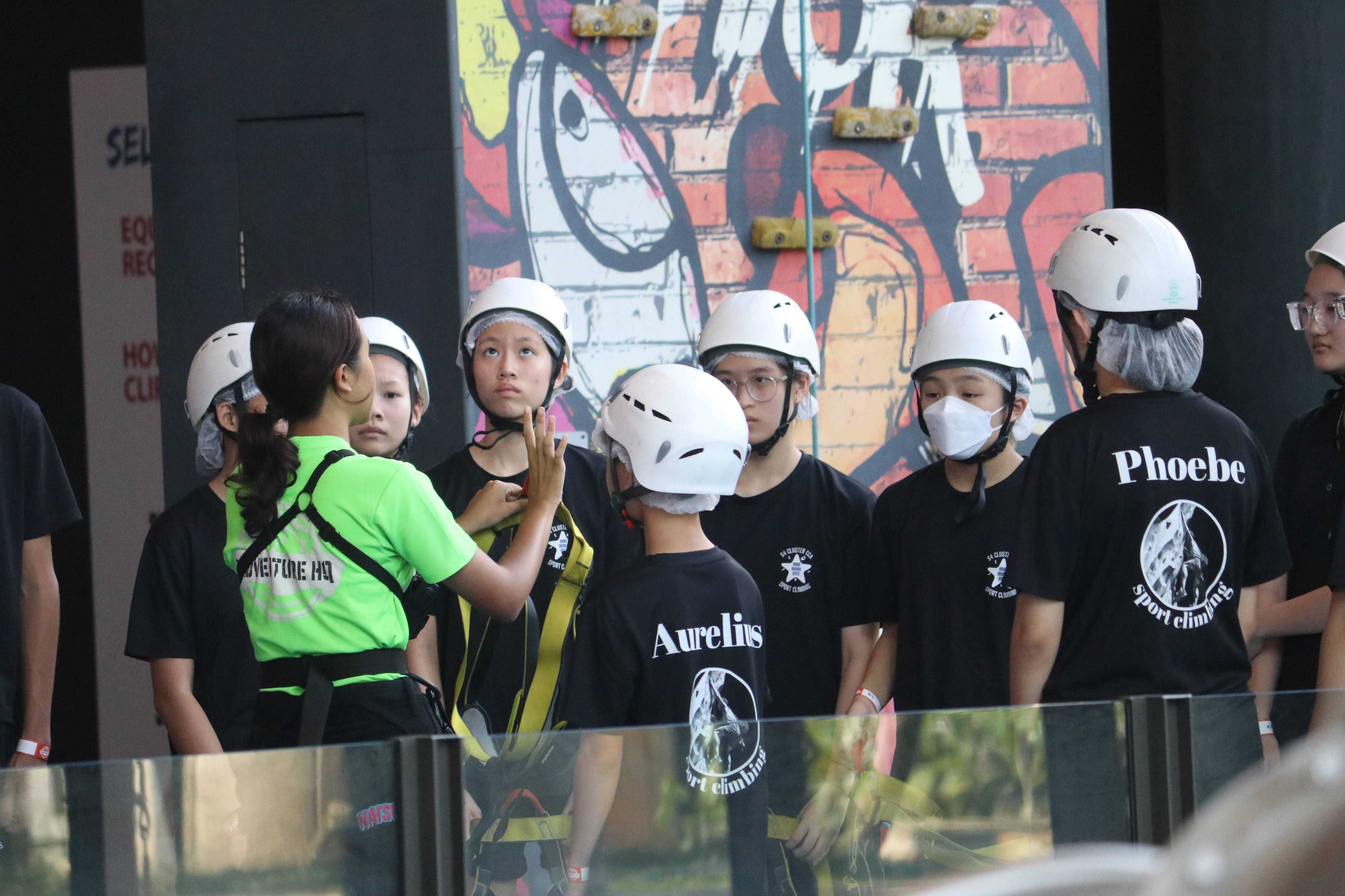 A group of young people in white helmets and black sport climbing shirts receive instructions from a guide in a green vest.